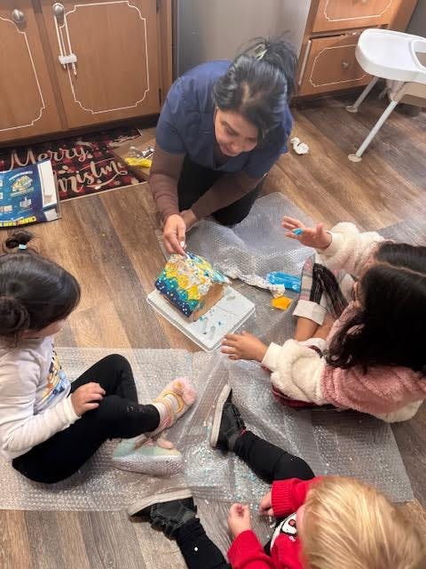 Person and children decorating a gingerbread house on the floor