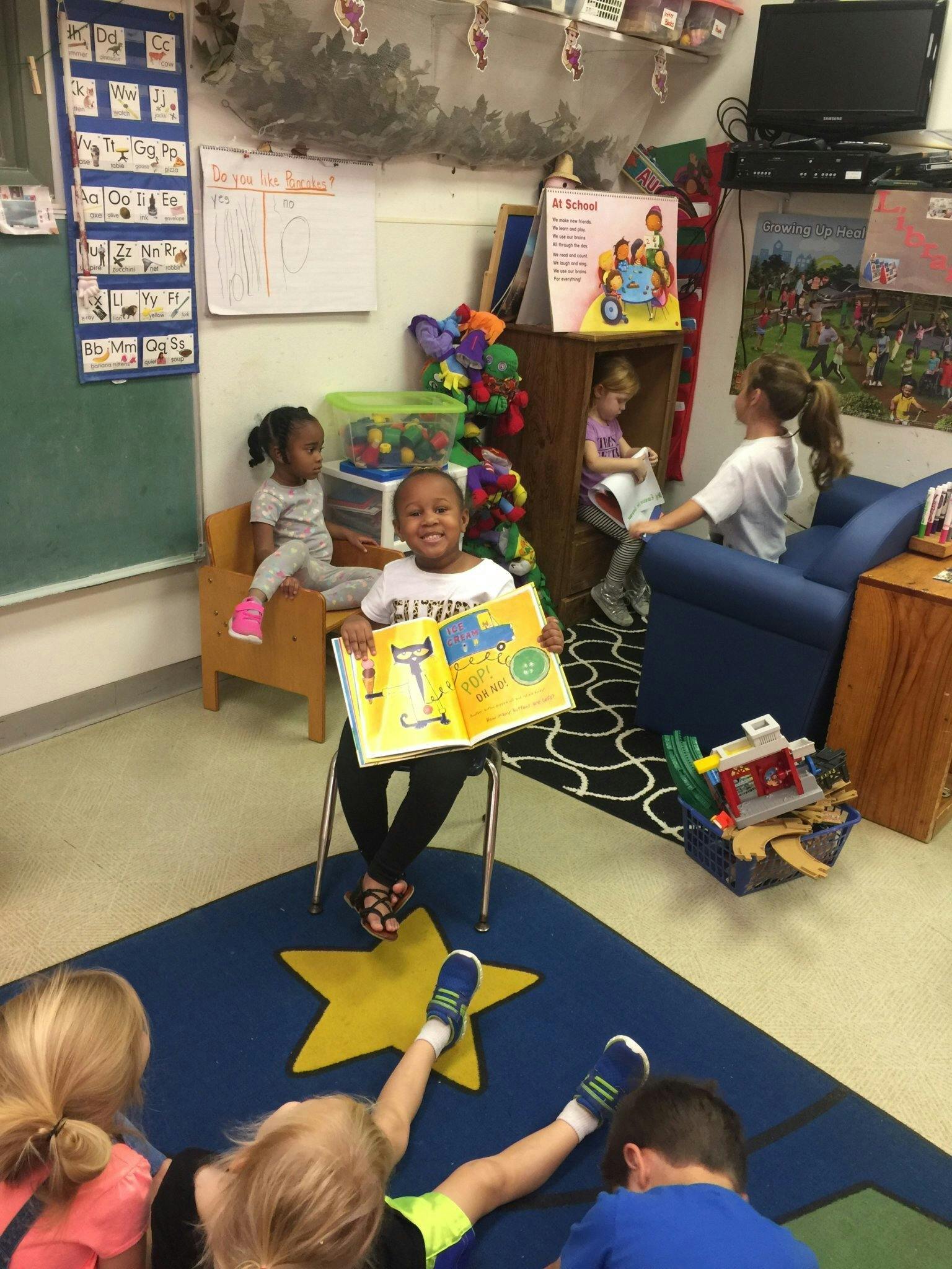 Child sitting on a chair holding an open book, other children on the floor