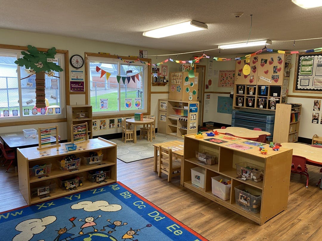 Classroom with tables, chairs, shelving units with clear bins, and a colorful rug