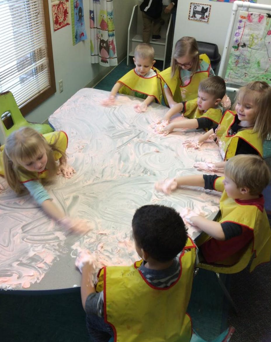 Children in yellow smocks playing with a white and pink substance on a table