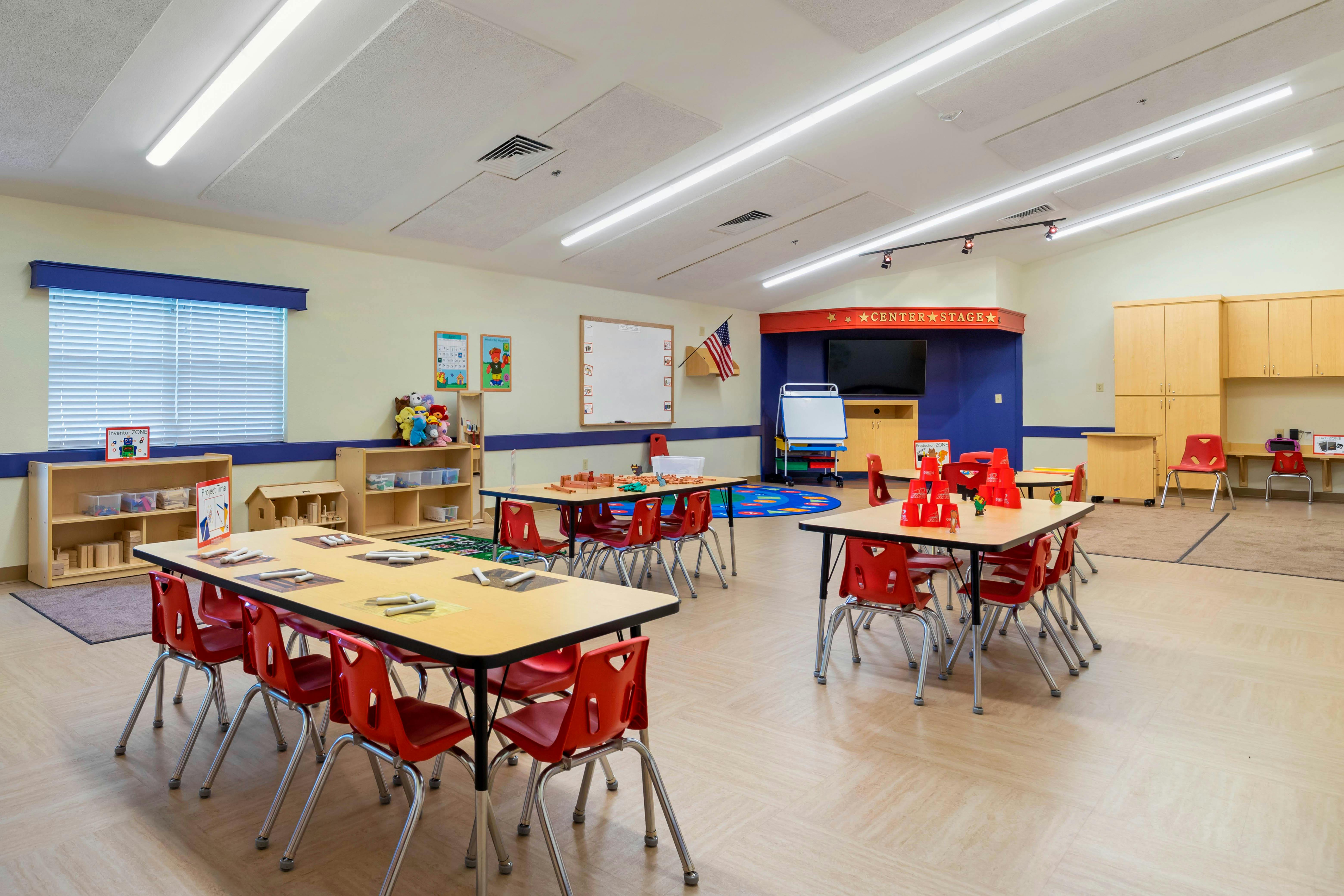 Empty classroom with small tables and red chairs, toys, and activity areas
