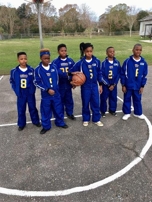 Six children in blue tracksuits on a basketball court, one holding a basketball
