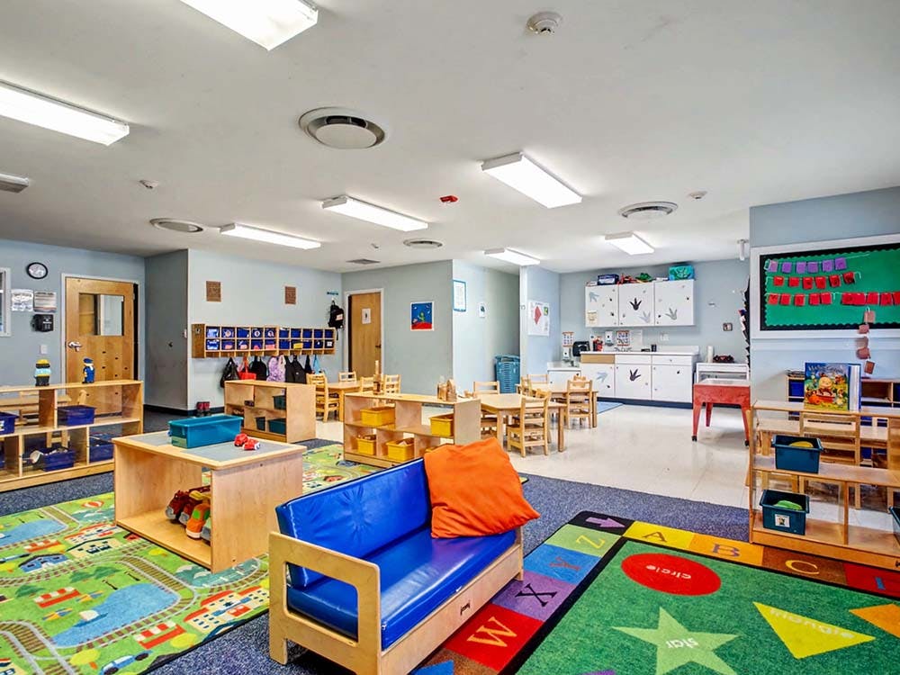 An empty children's classroom with small tables, chairs, and play mats