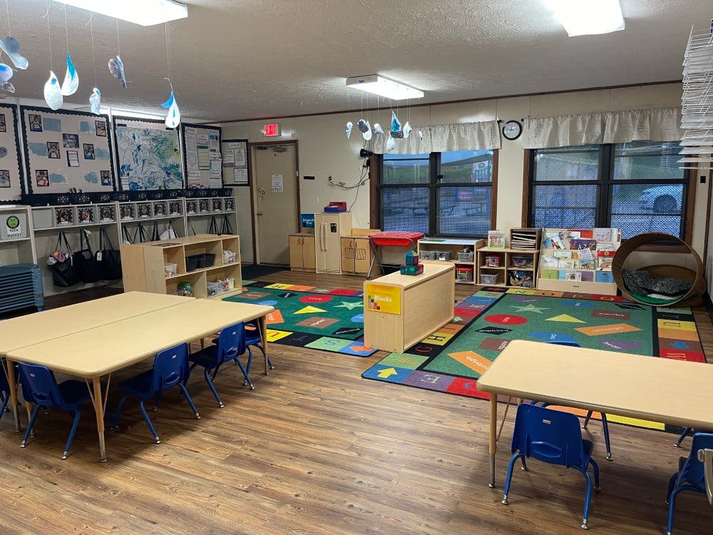 Classroom with child-sized tables and chairs, shelves with toys, and colorful rugs