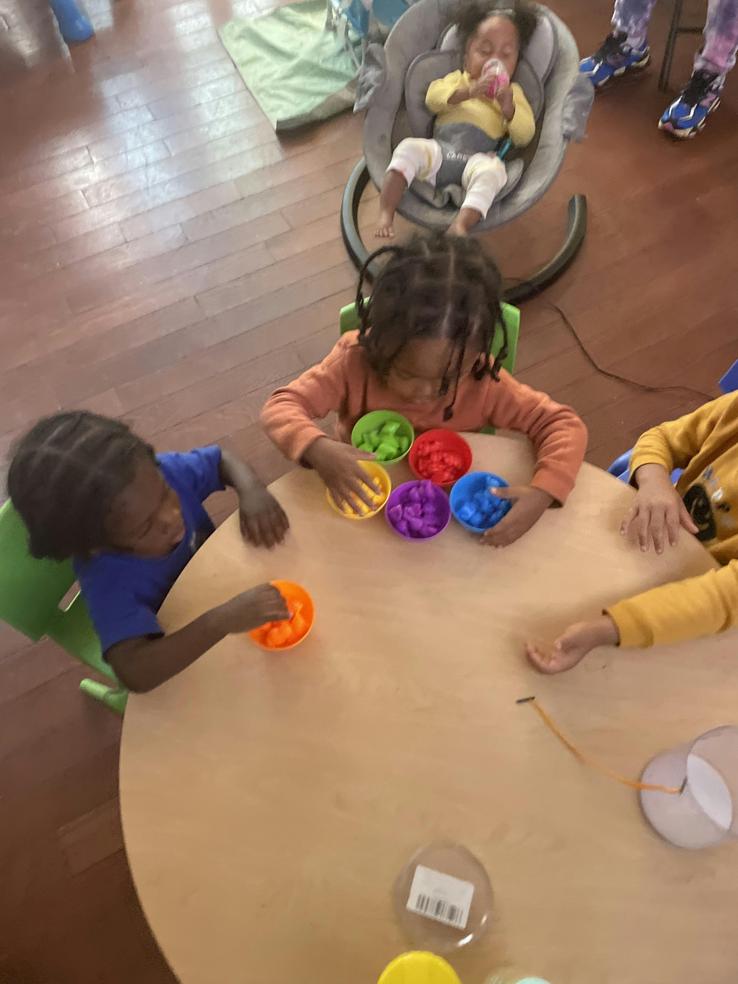 Children playing with colorful bowls at a table, one child in a baby bouncer