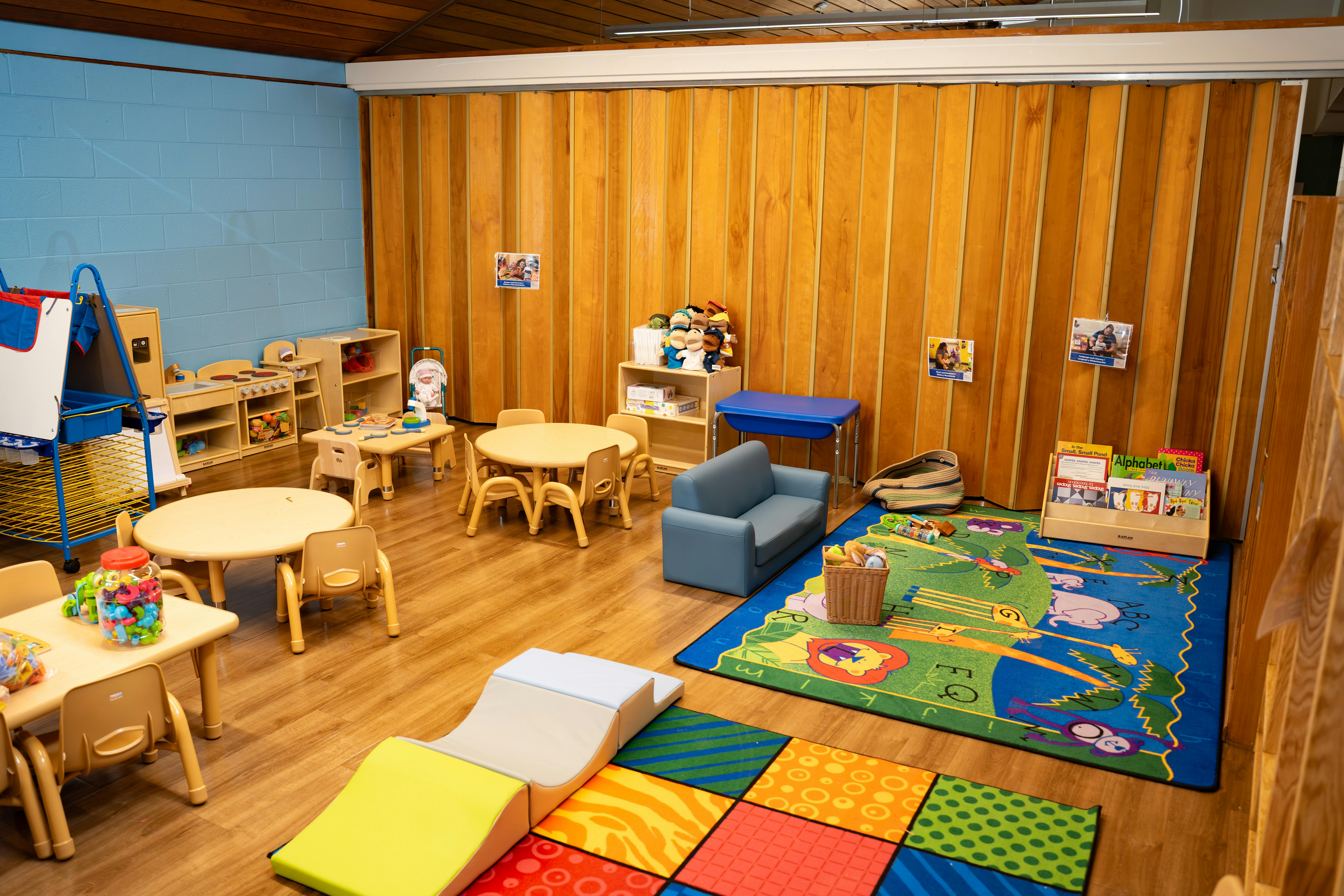 Empty preschool classroom with small tables, chairs, play kitchen, and colorful rugs