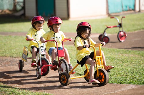 Three children wearing helmets riding tricycles outdoors