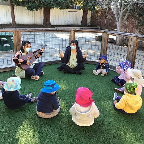 People and children sitting in a circle on grass, one person playing a ukulele