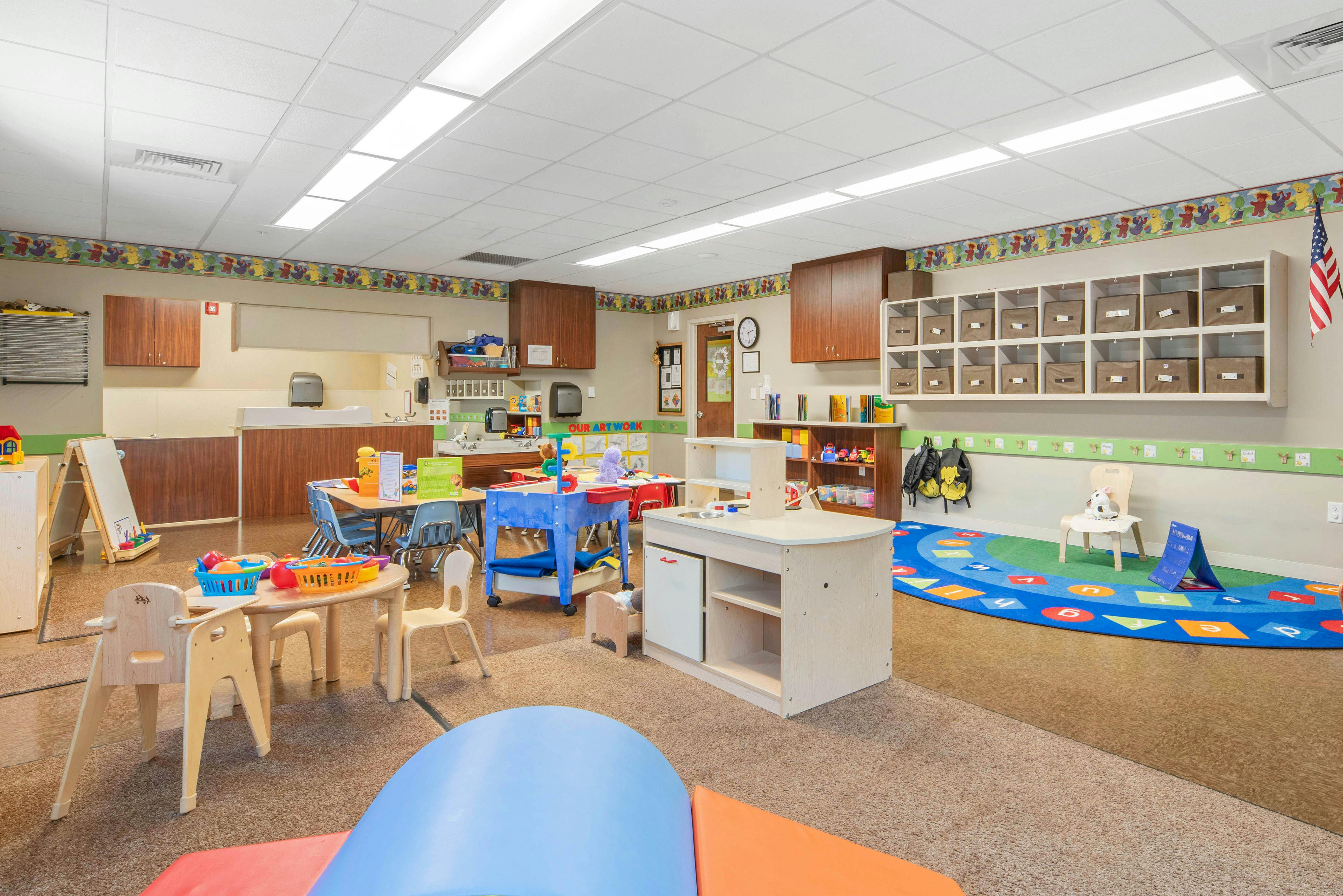 Classroom filled with small tables, chairs, play kitchen, and a colorful rug