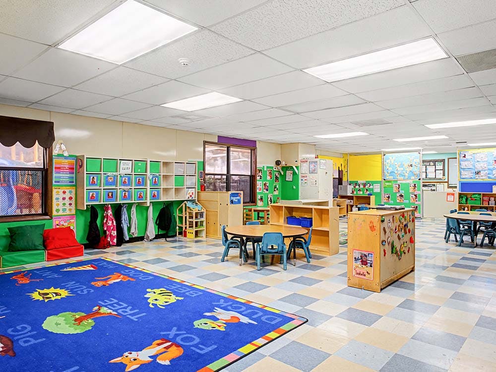 A brightly lit classroom with a blue animal rug and small tables and chairs