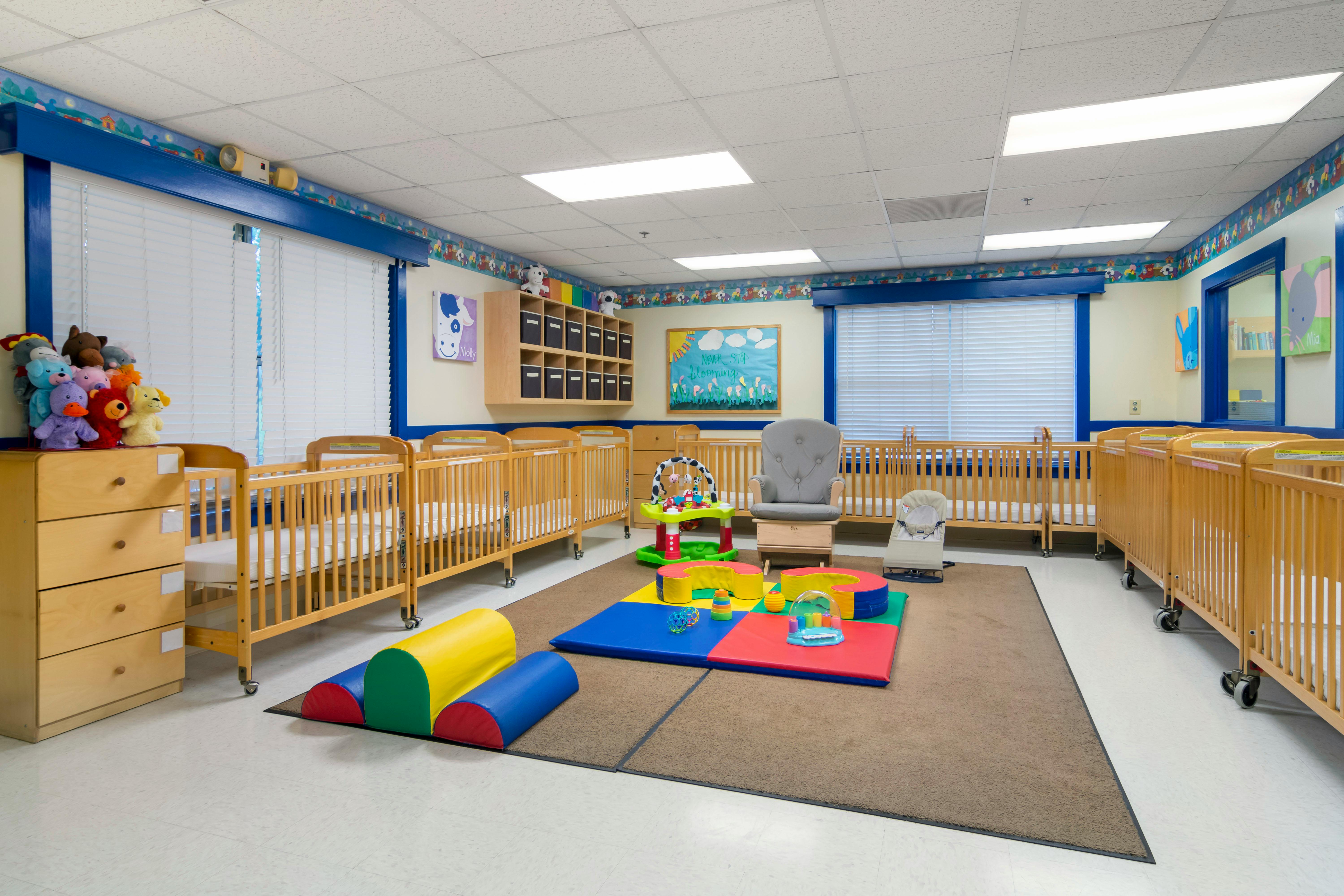 A brightly lit nursery with multiple wooden cribs, playmats, and a rocking chair
