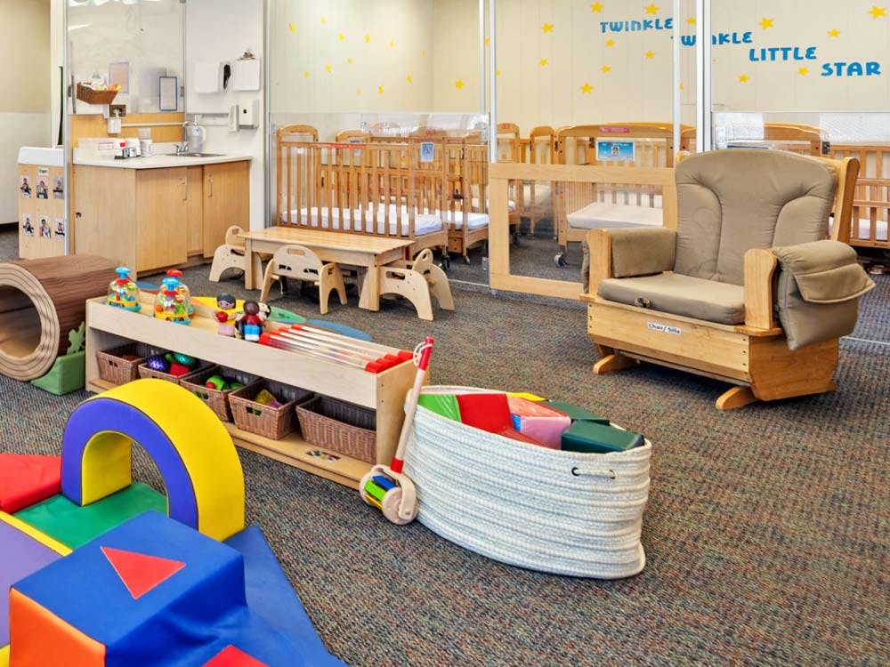 Childcare room with play mats, wooden toys, cribs, and a rocking chair