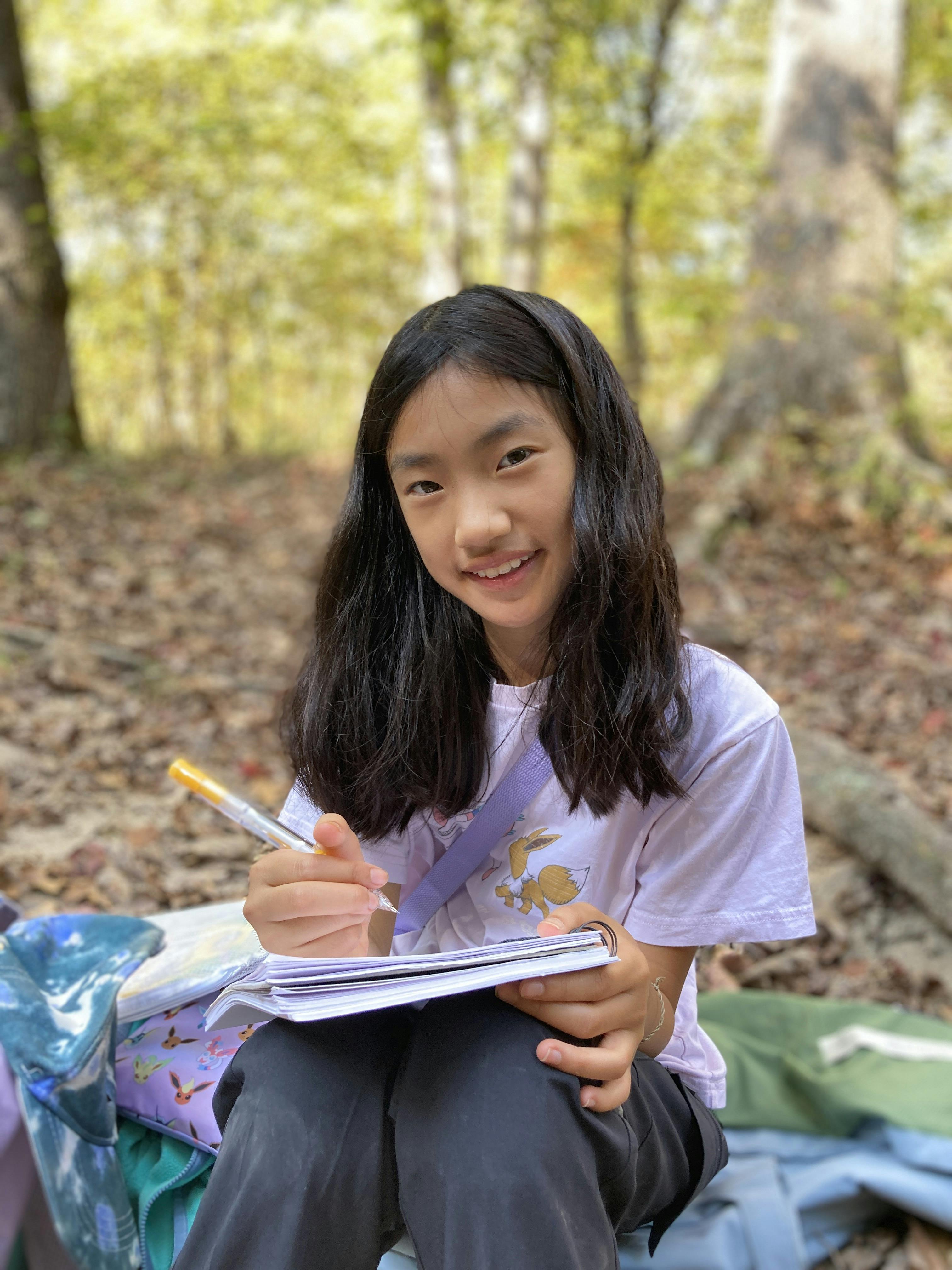 Child writing in a notebook while sitting in a wooded area