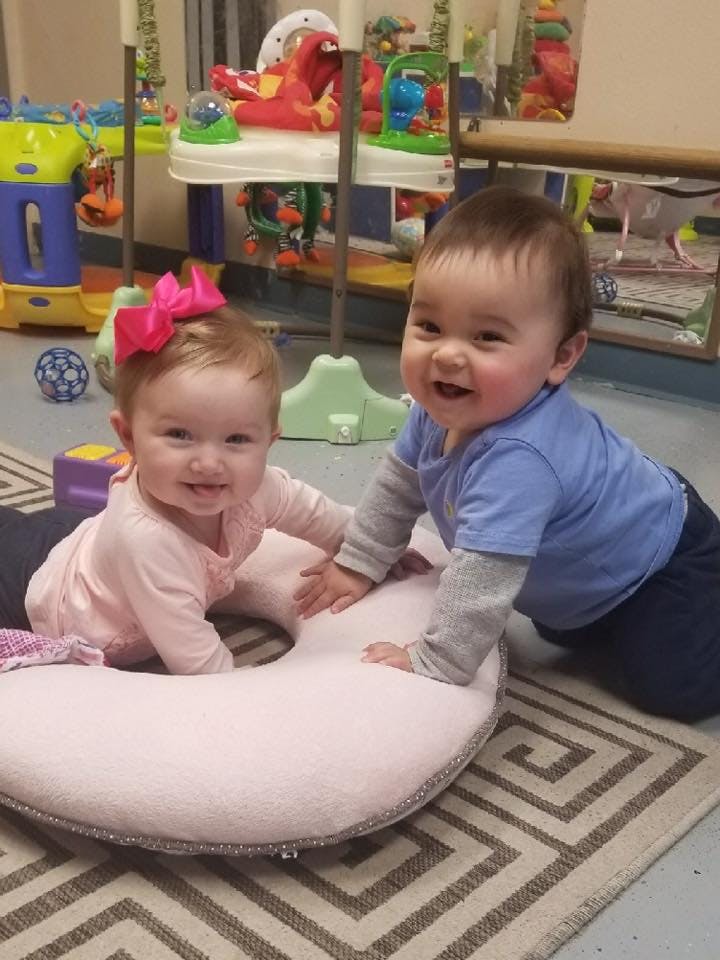 Two babies on a rug, one on a pink nursing pillow