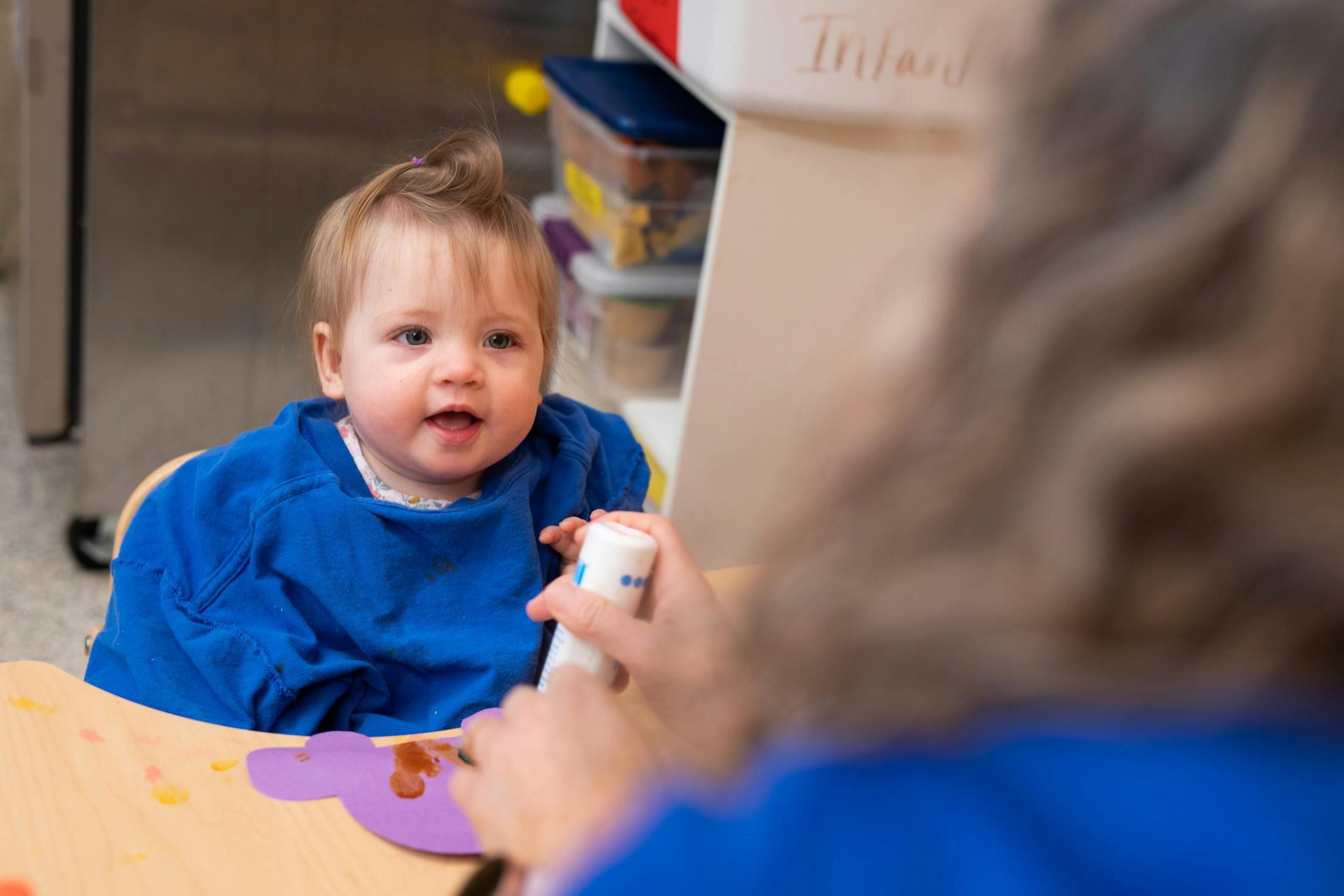Child in a high chair looking at a person holding a bottle
