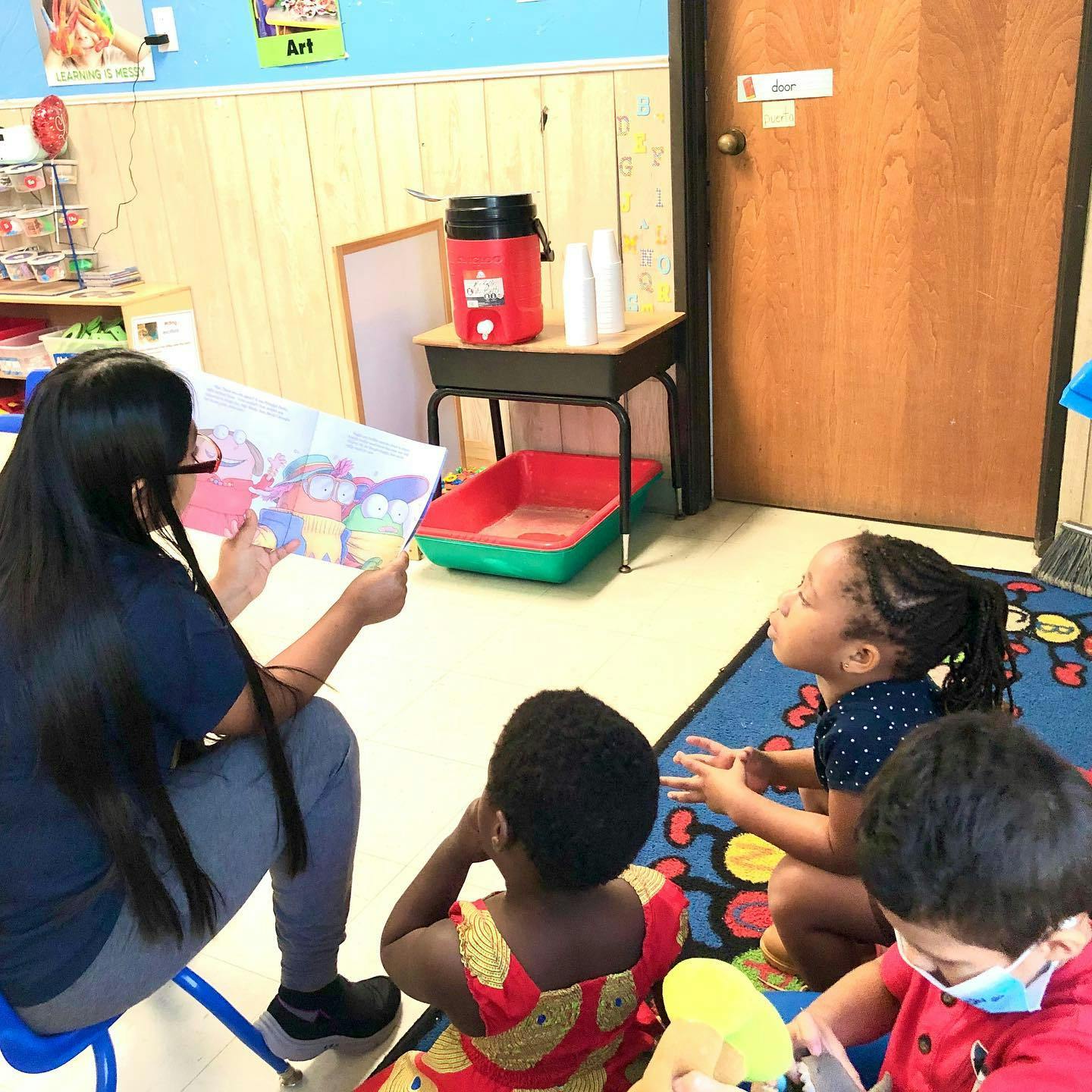 Person reading a book to three children in a classroom