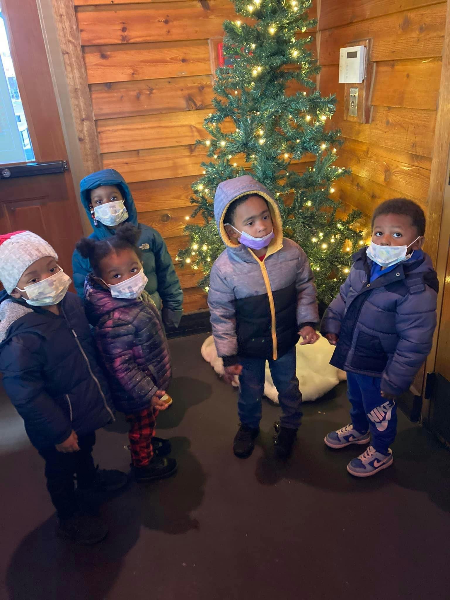 Four children wearing face masks and winter coats standing near a Christmas tree