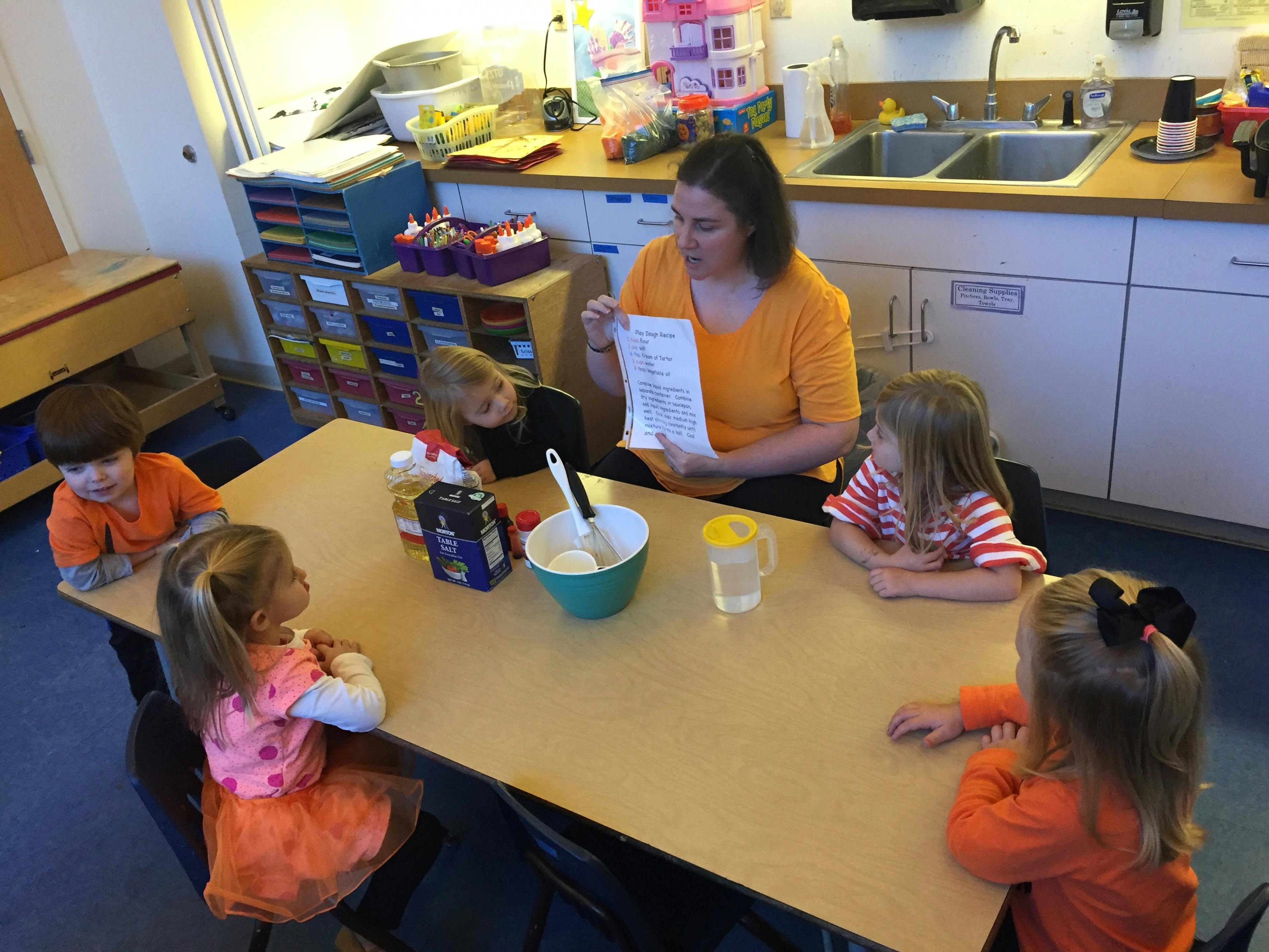 Person reading to four children at a table with a bowl and pitcher