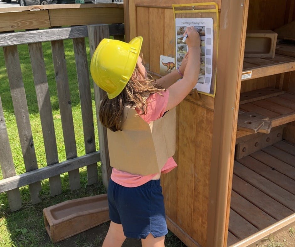 Child in a yellow hard hat and cardboard vest reaching for a paper on a wooden structure