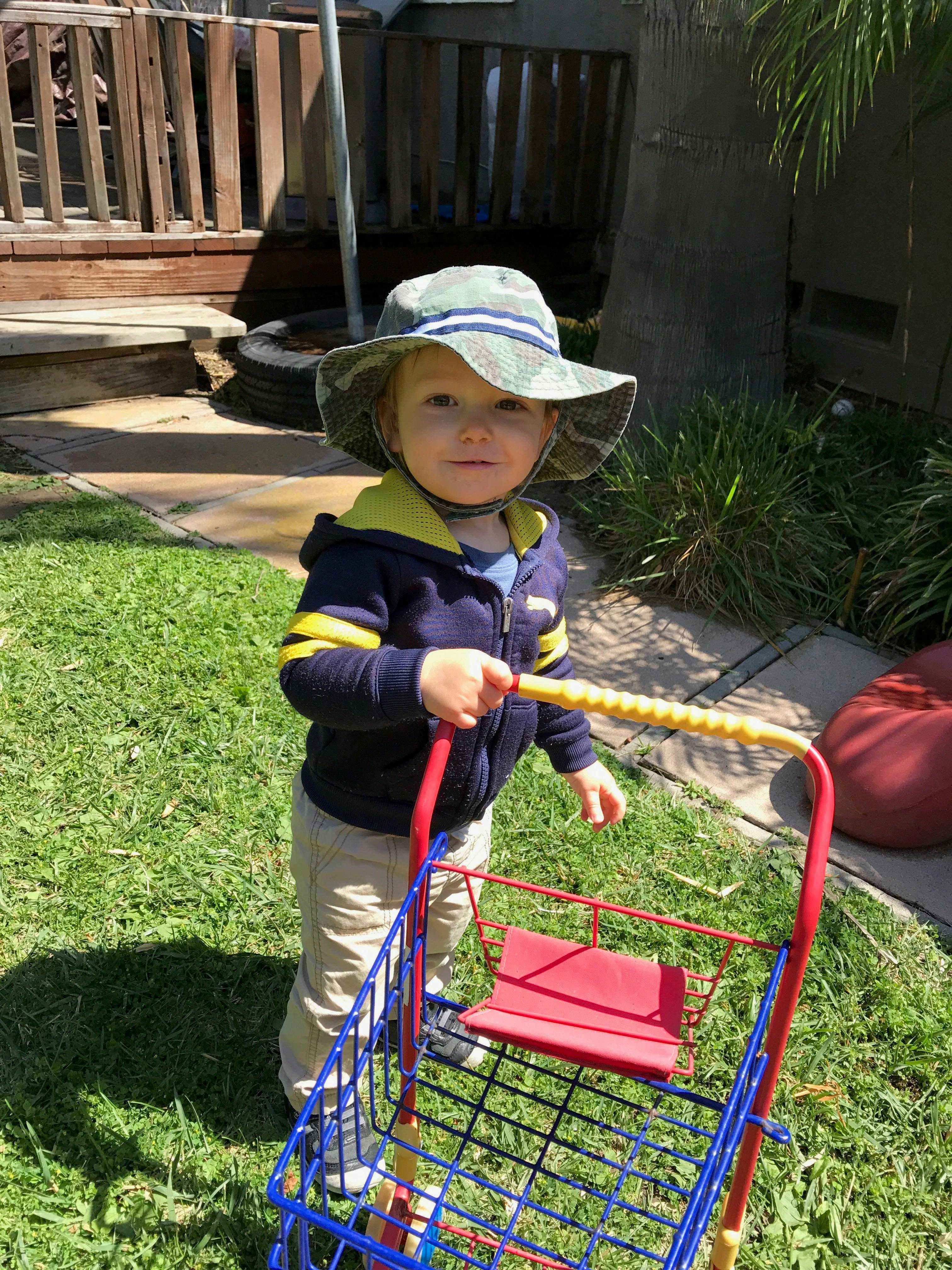 Child wearing a bucket hat pushing a toy shopping cart in a yard