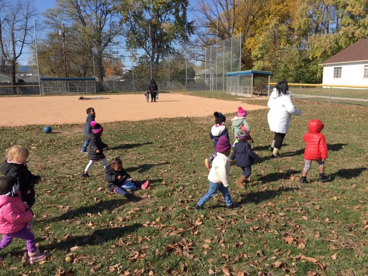 Children and adults on a grassy field next to a baseball diamond