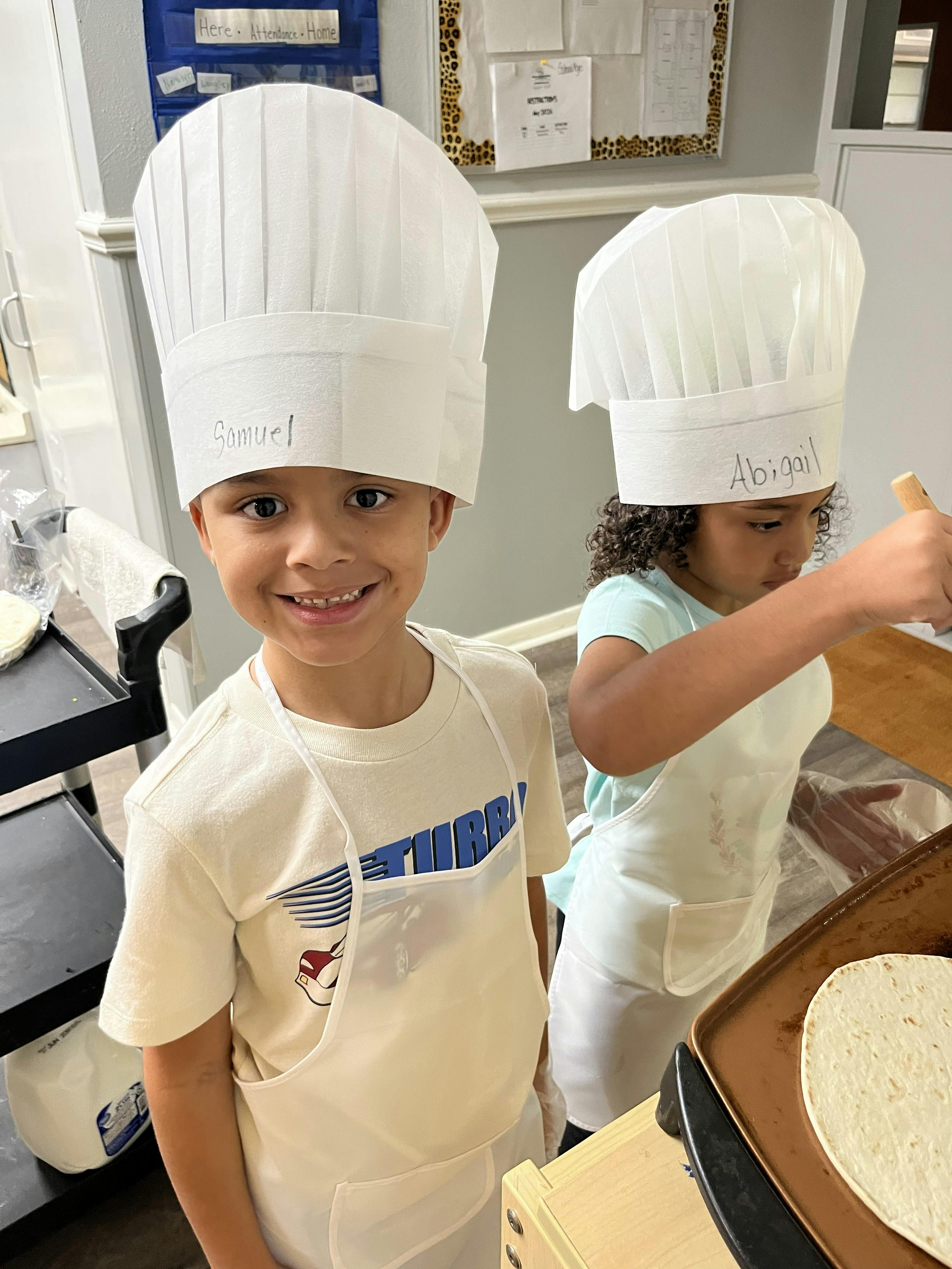 Two children wearing chef hats and aprons preparing food