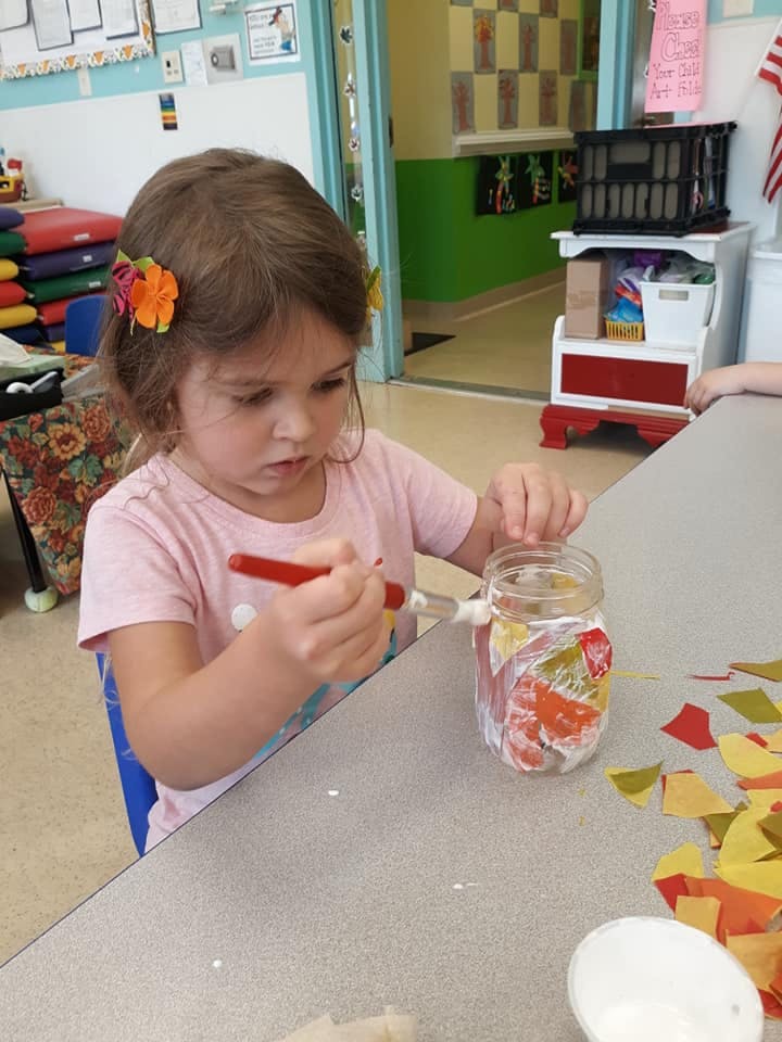 Child painting a glass jar with colorful paper cutouts