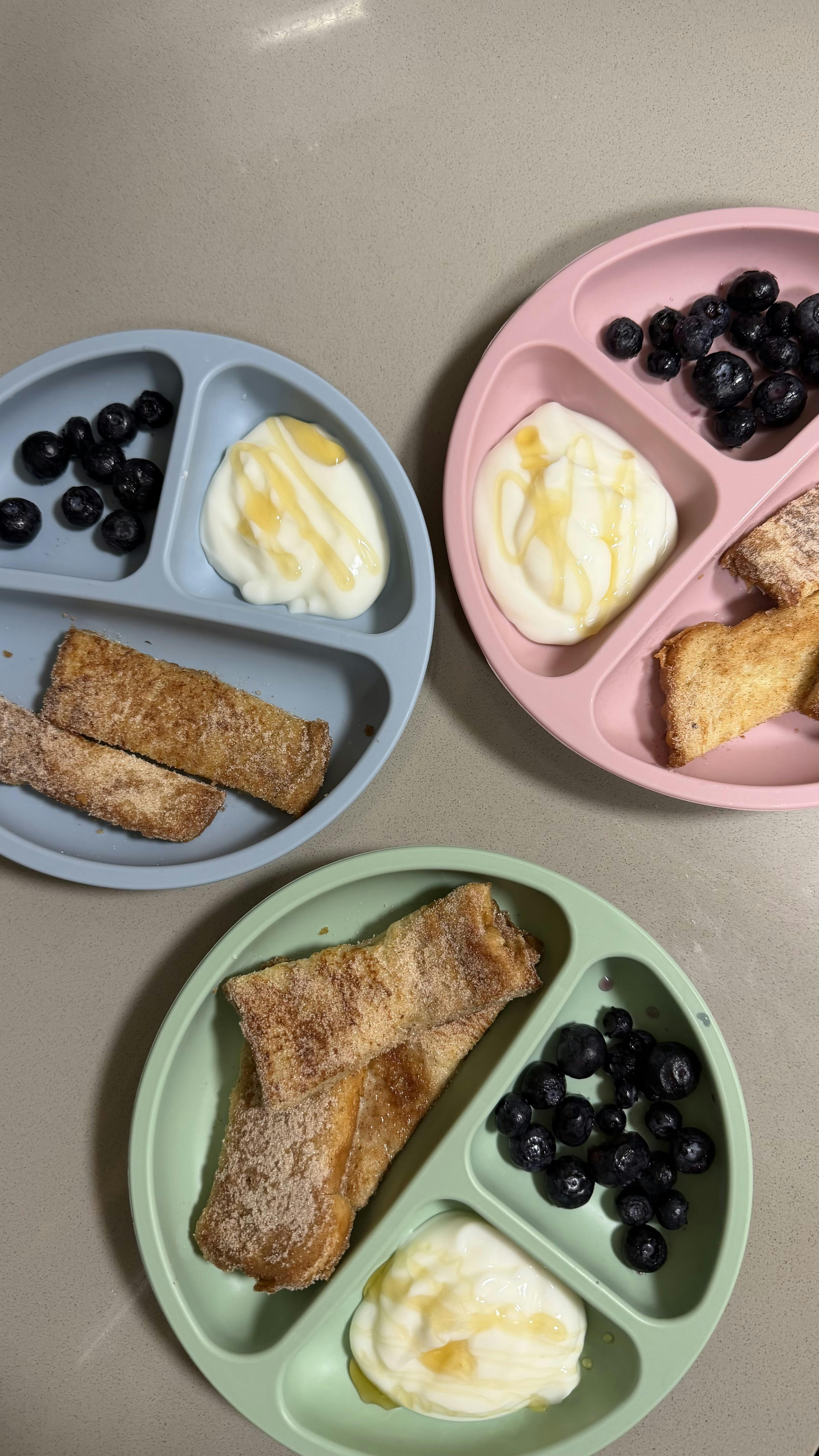 Three divided plates containing french toast sticks, yogurt, and blueberries