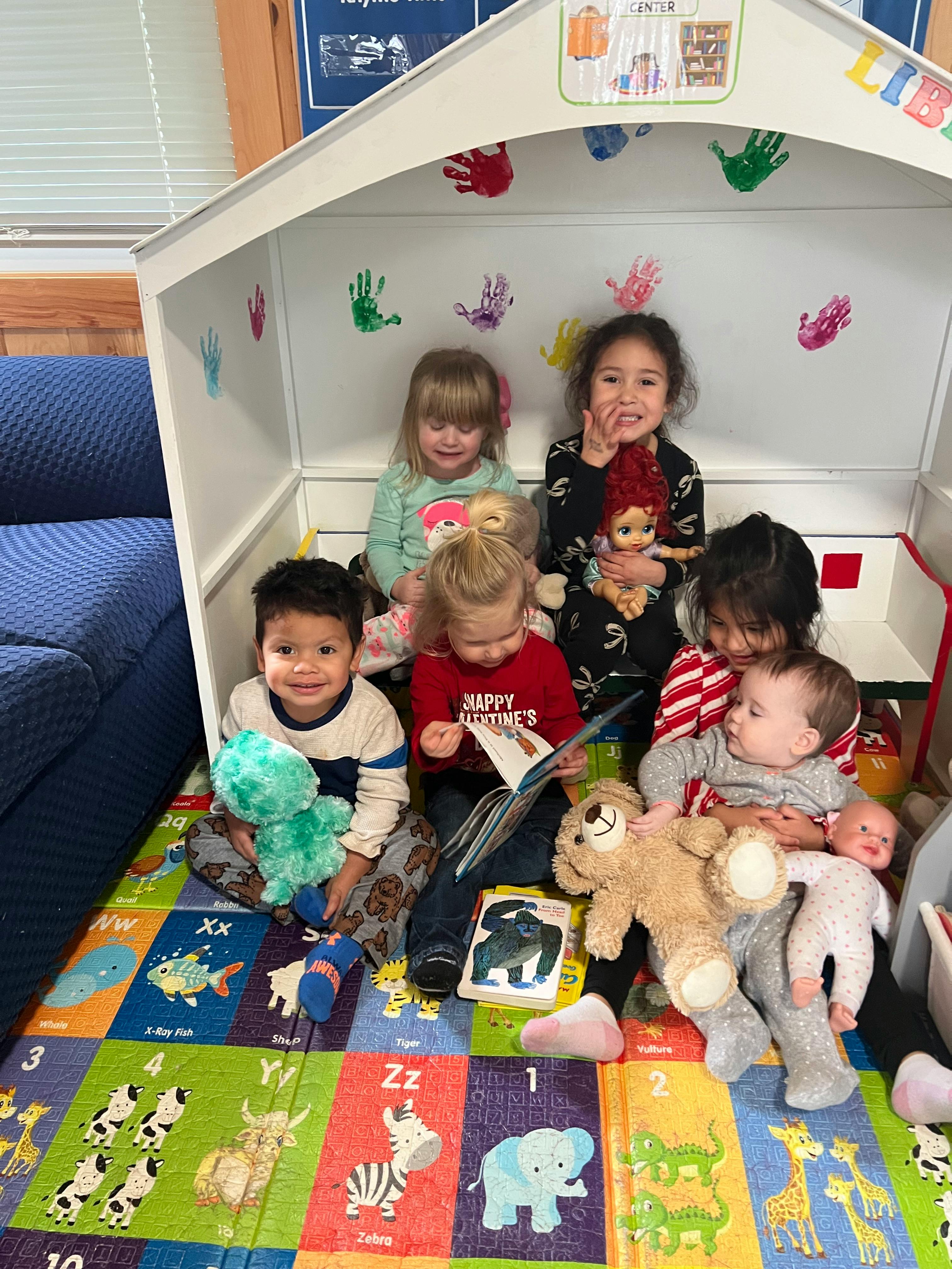 Group of children and babies sitting in a playhouse with stuffed animals and baby dolls