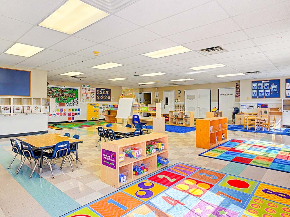 Empty classroom with small tables and chairs, colorful rugs, and shelves of toys