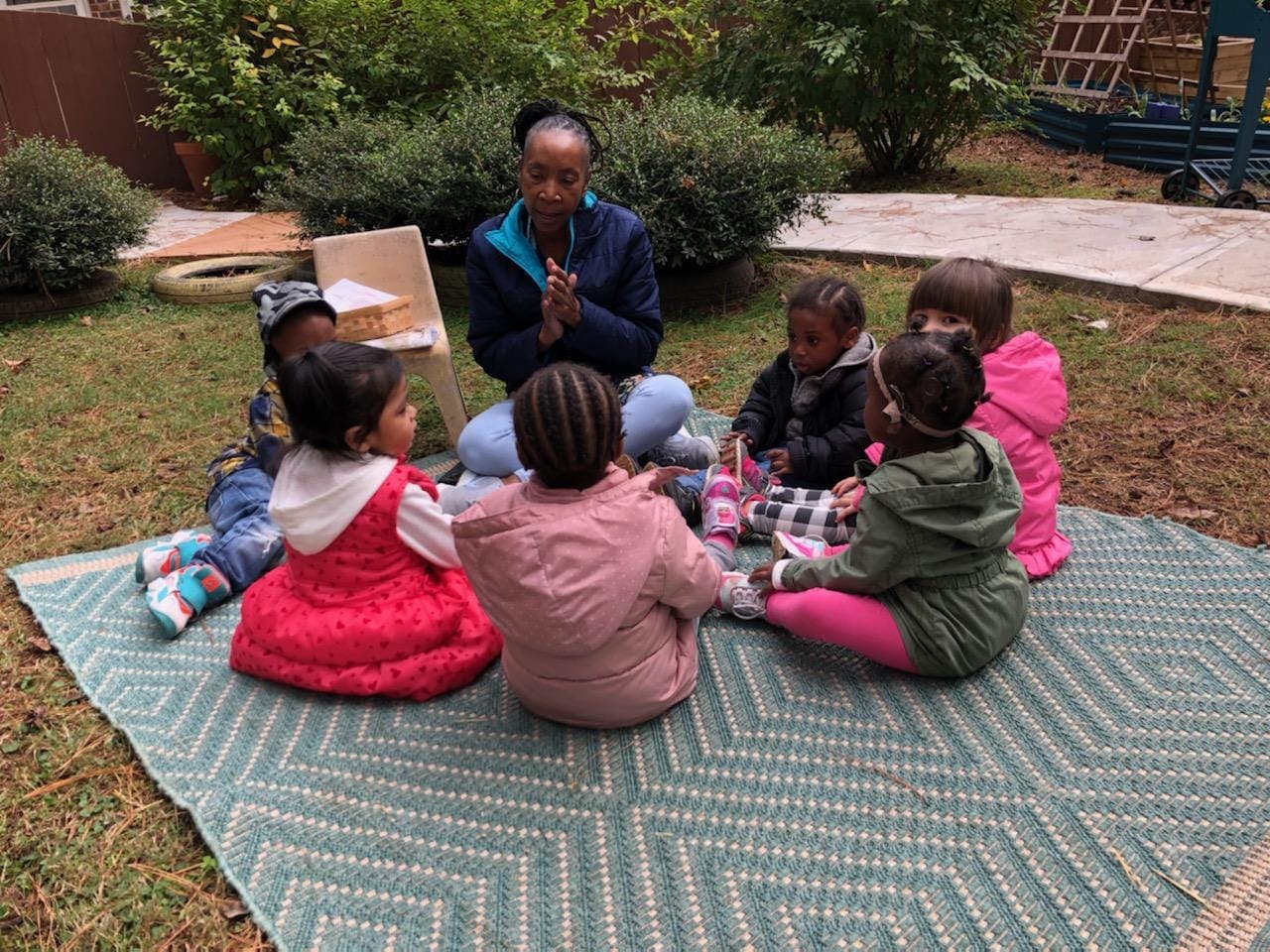 Adult and multiple children sitting in a circle on a mat outdoors
