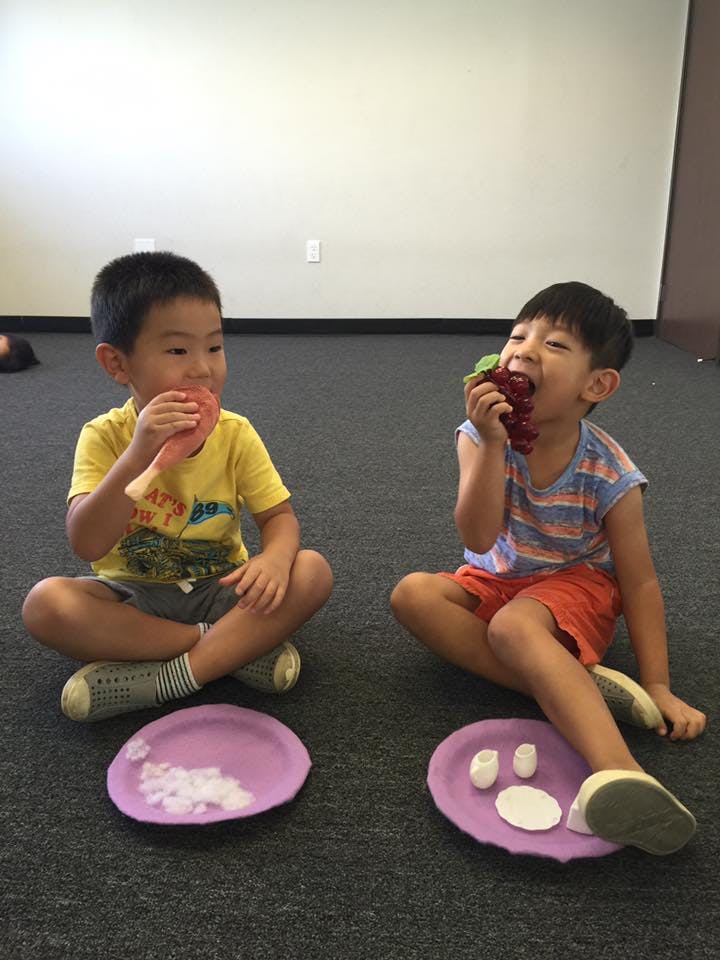 Two children sitting on the floor, one eating a red fruit, the other eating grapes
