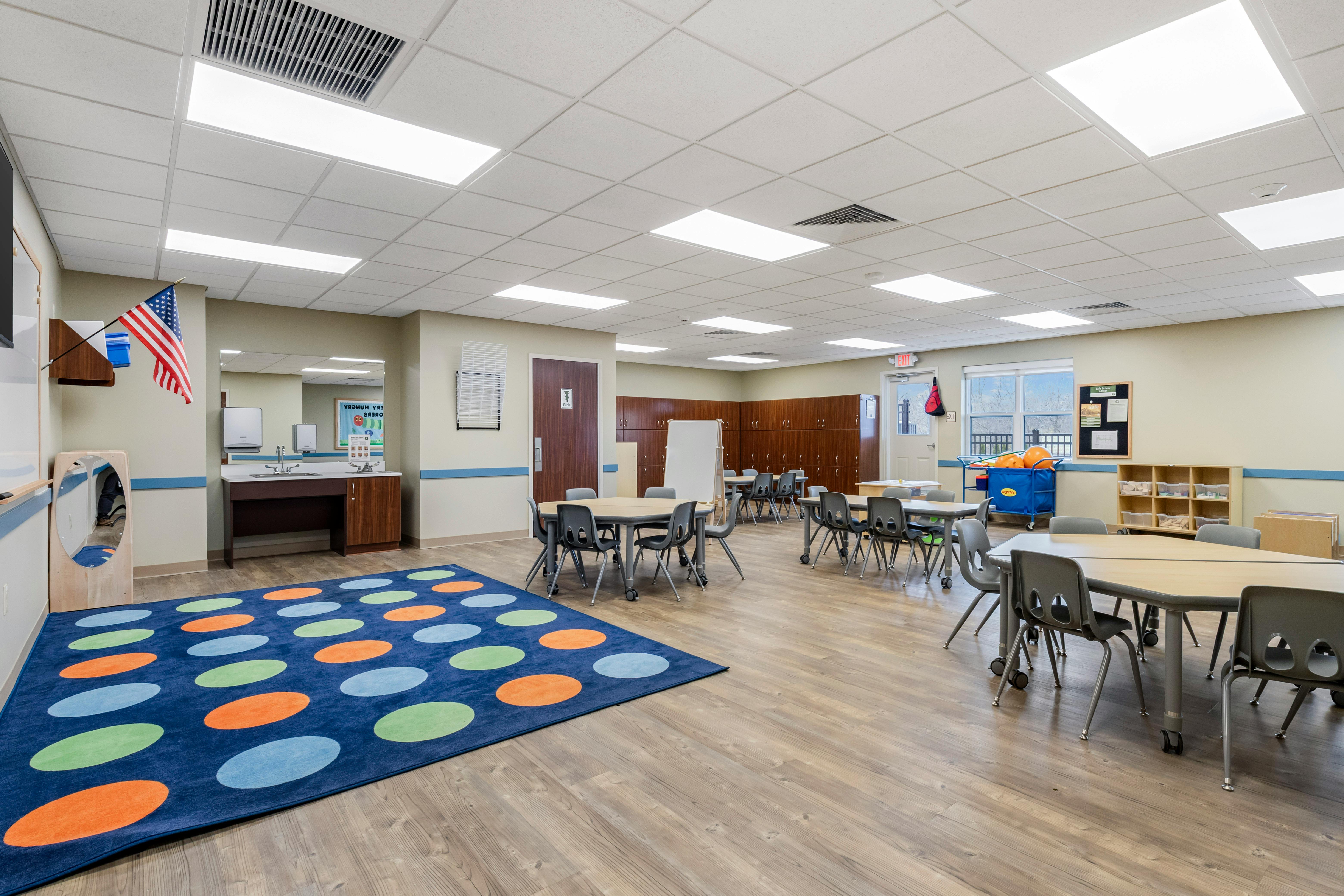 Classroom with small tables and chairs, wood-patterned floor, and a colorful rug