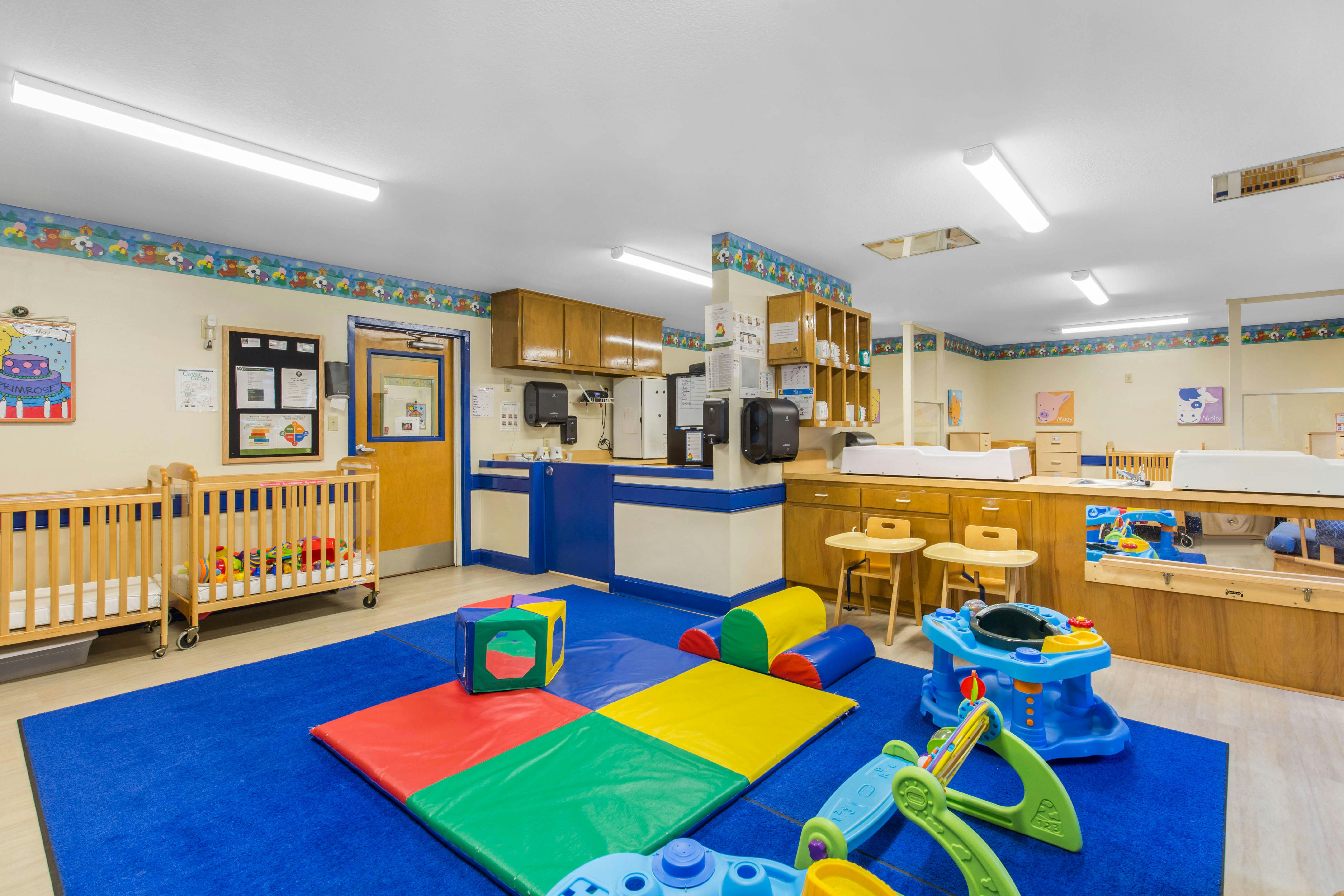 A colorful childcare classroom with cribs, play mats, and activity stations