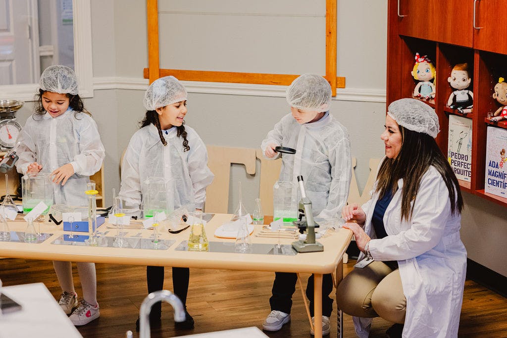 Adult and three children in lab coats and hairnets at a table with science equipment