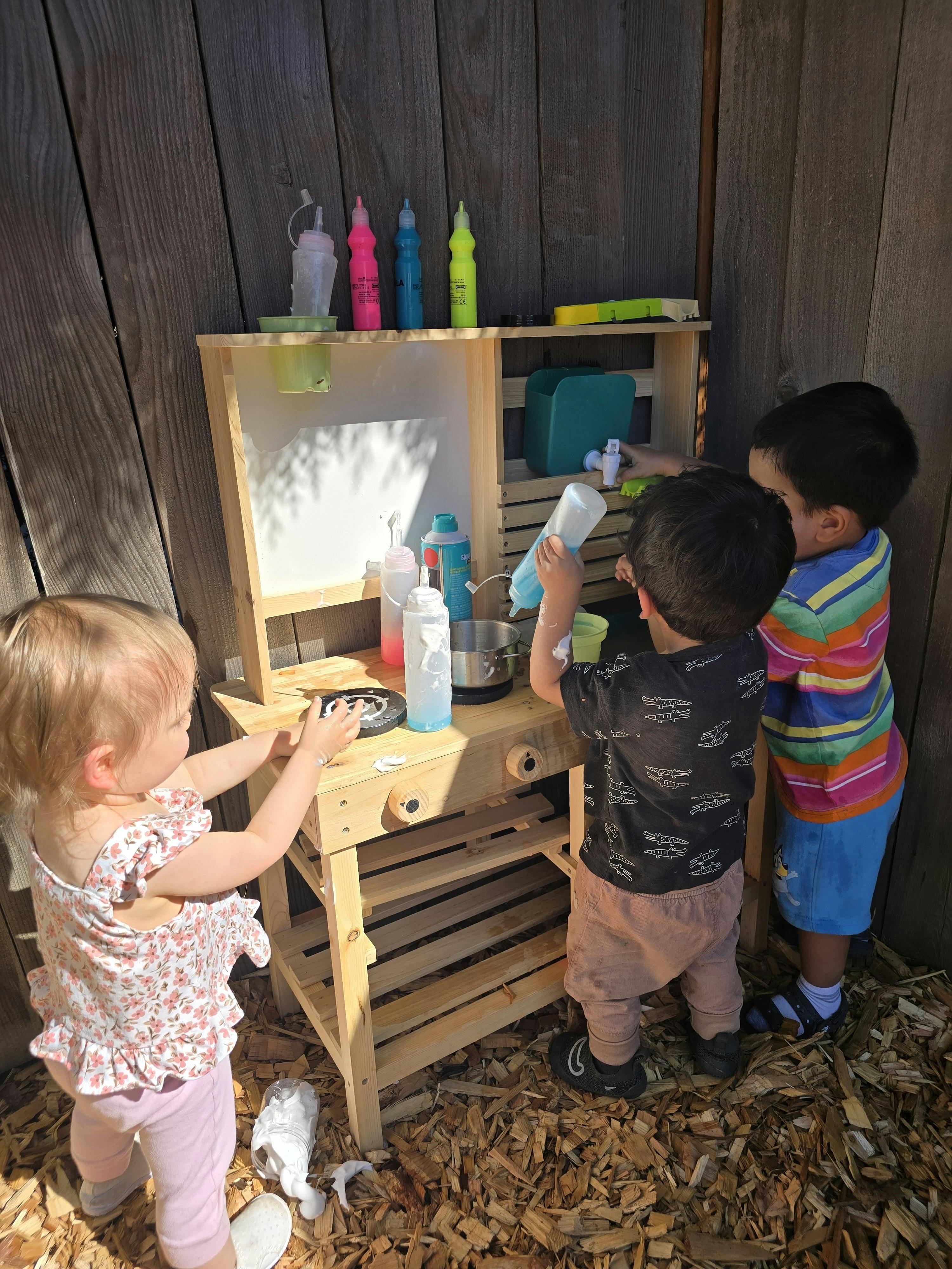 Three children playing at a wooden outdoor play kitchen