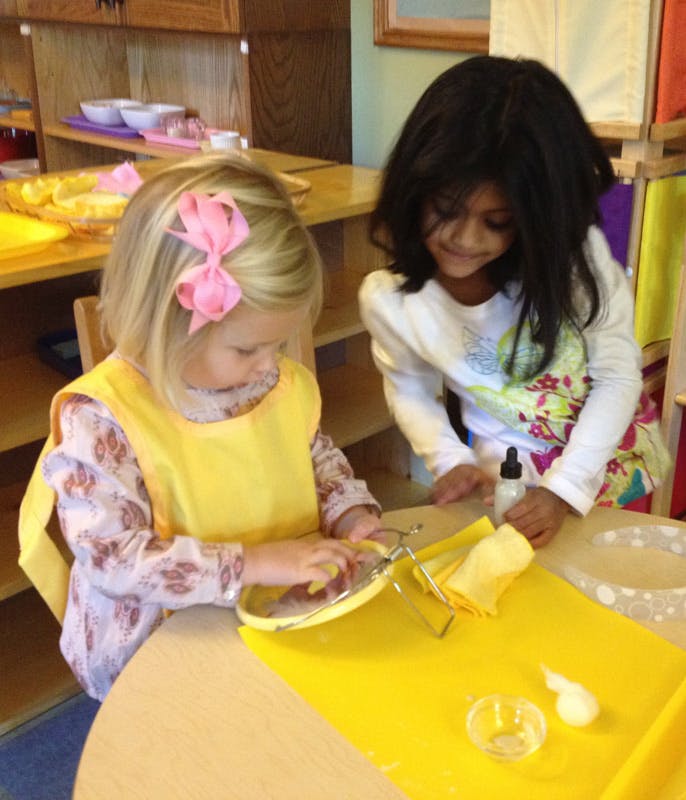 Two children at a table, one cleaning a dish, the other watching