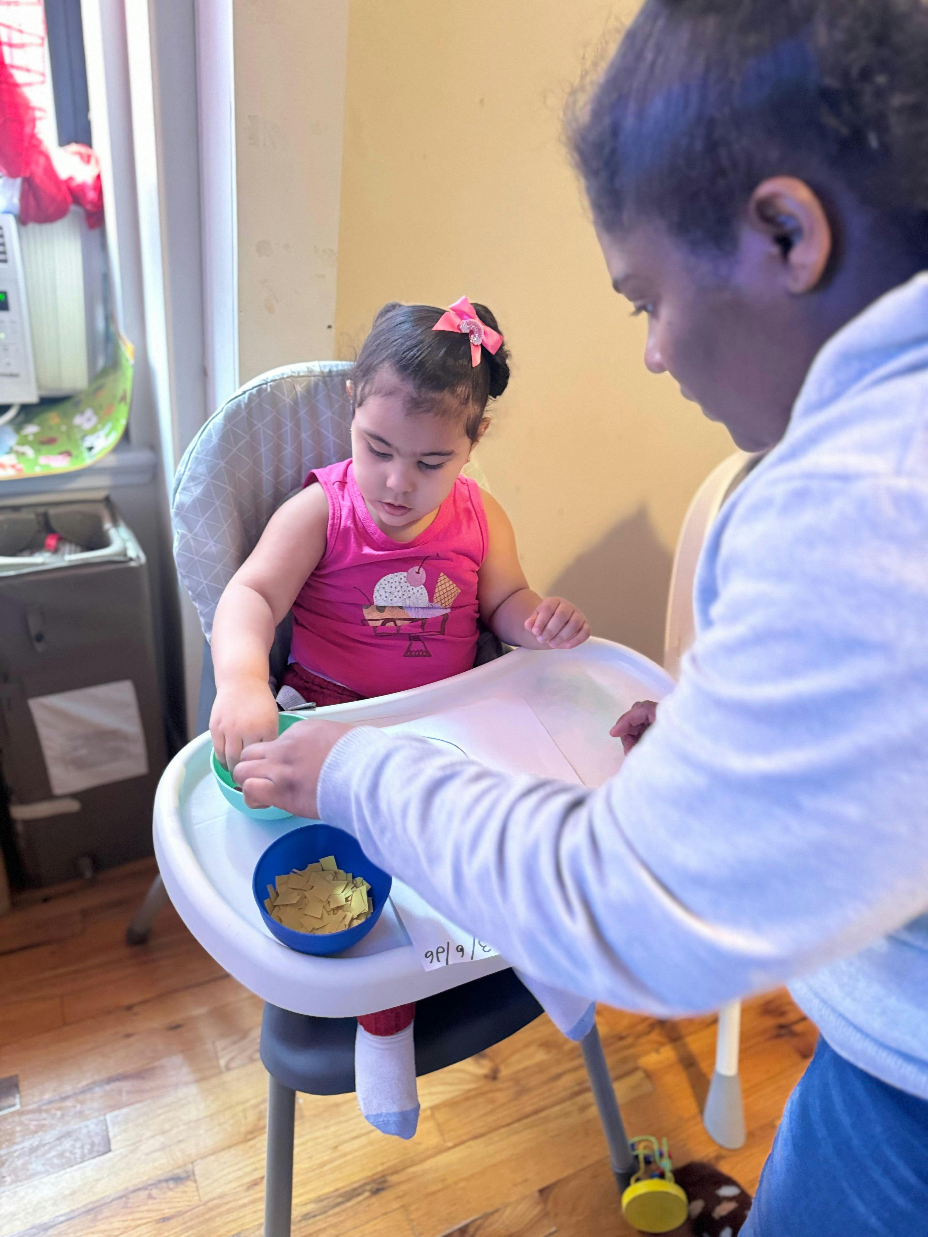 Child in high chair reaching for small bowl next to a person