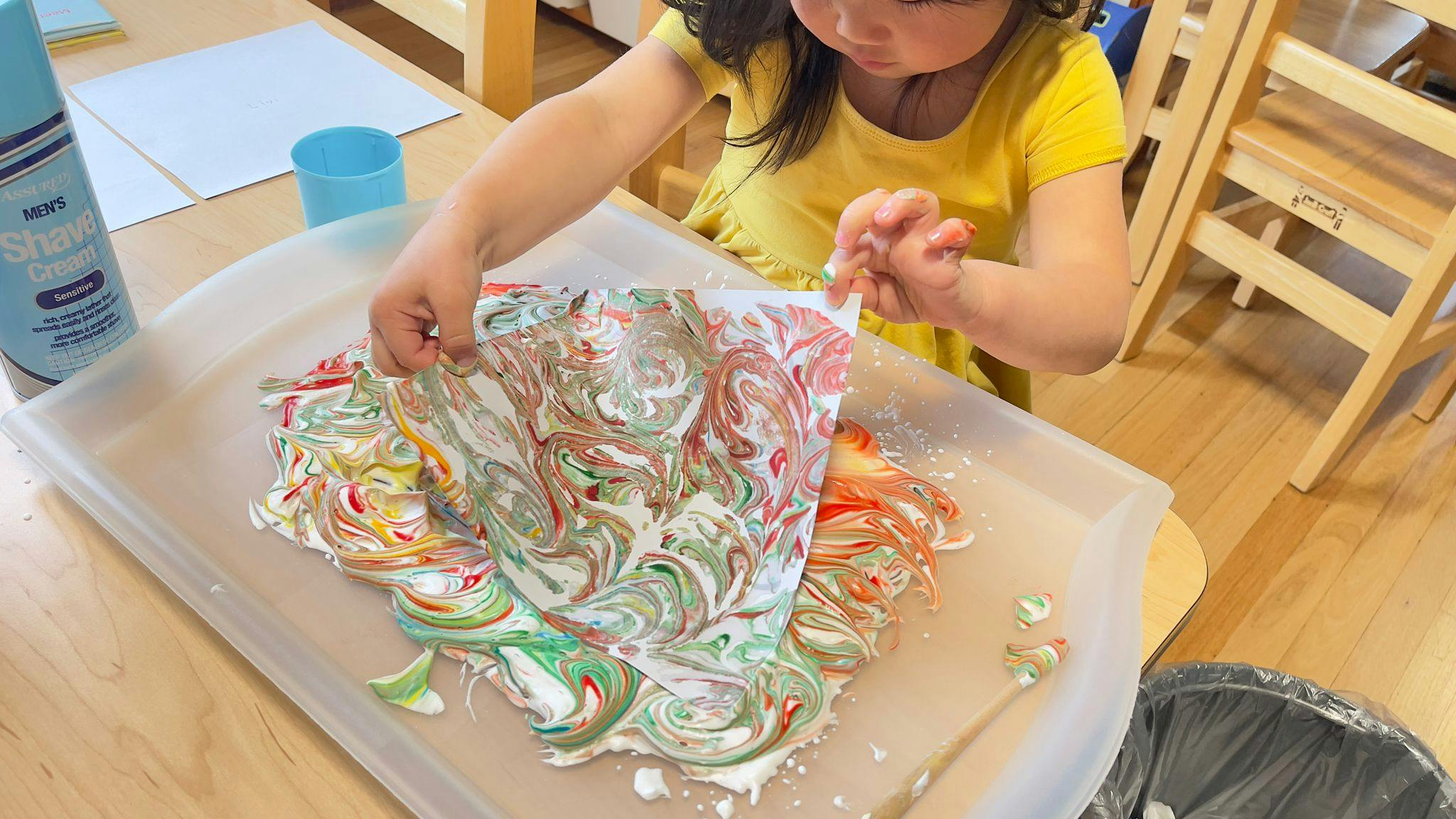 Child lifting paper from a tray of swirled shaving cream and paint
