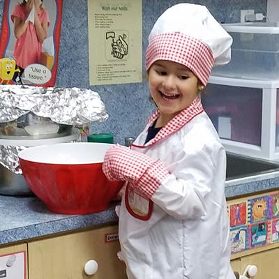 Child in chef's hat and coat holding a red mixing bowl