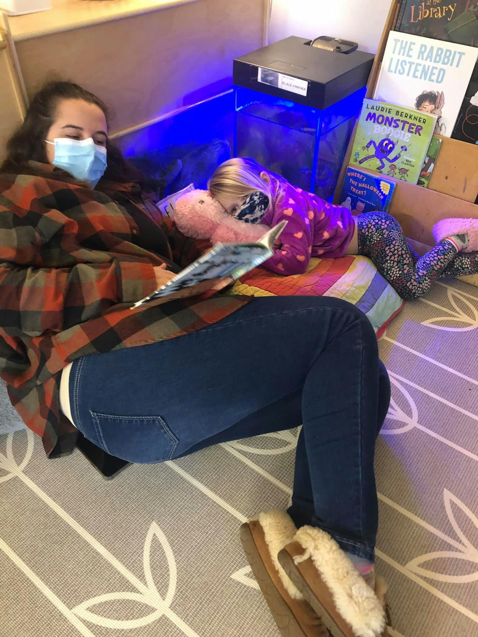 Person in mask reading book with child resting nearby