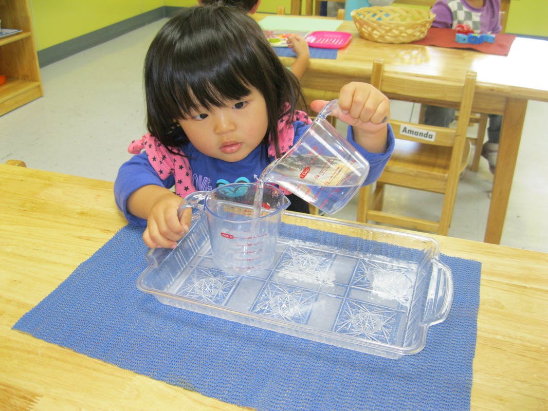 Child pouring water from a measuring cup into a clear container in a classroom