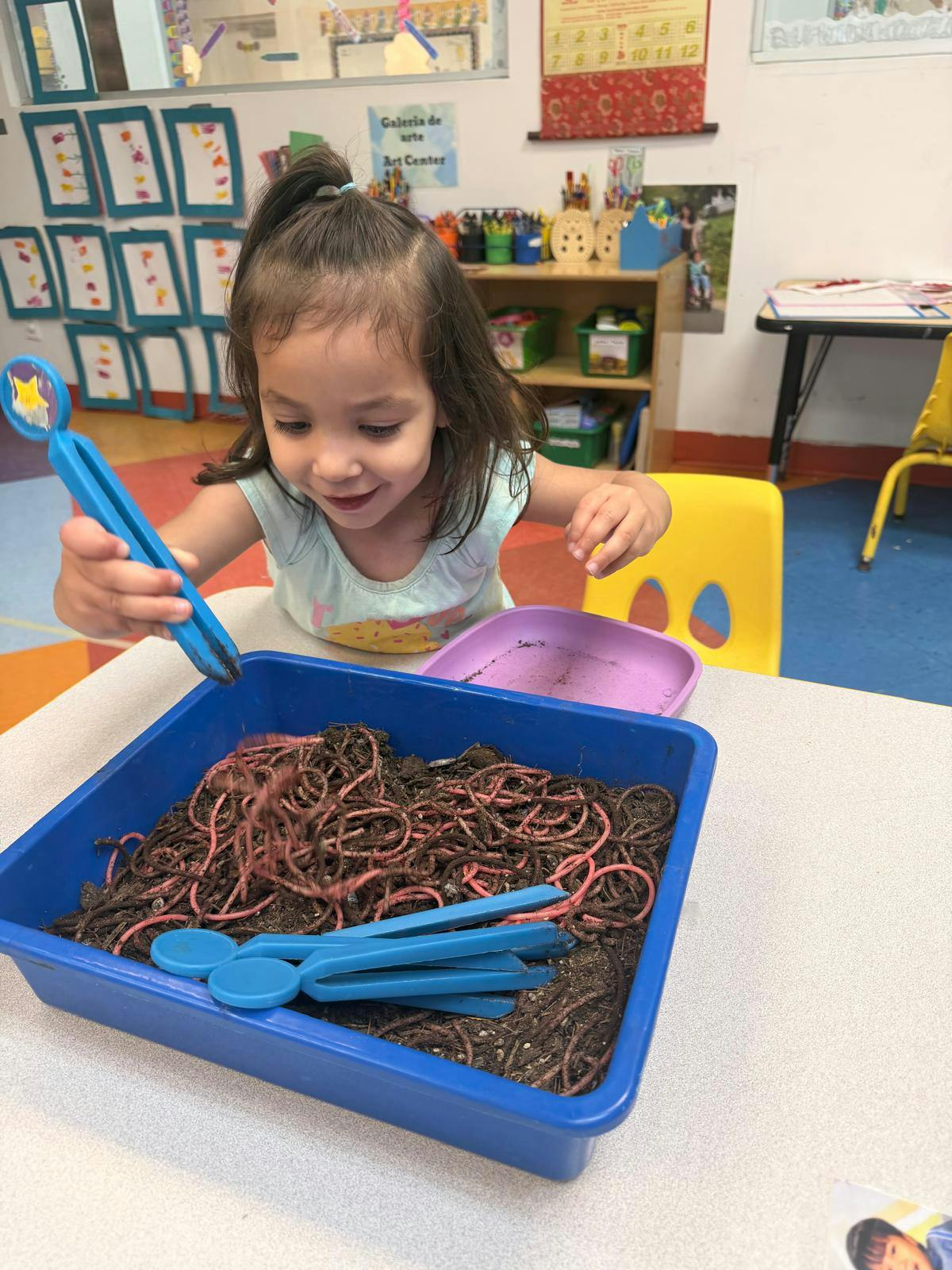 Child using plastic tongs to pick up faux worms from a tray of dirt