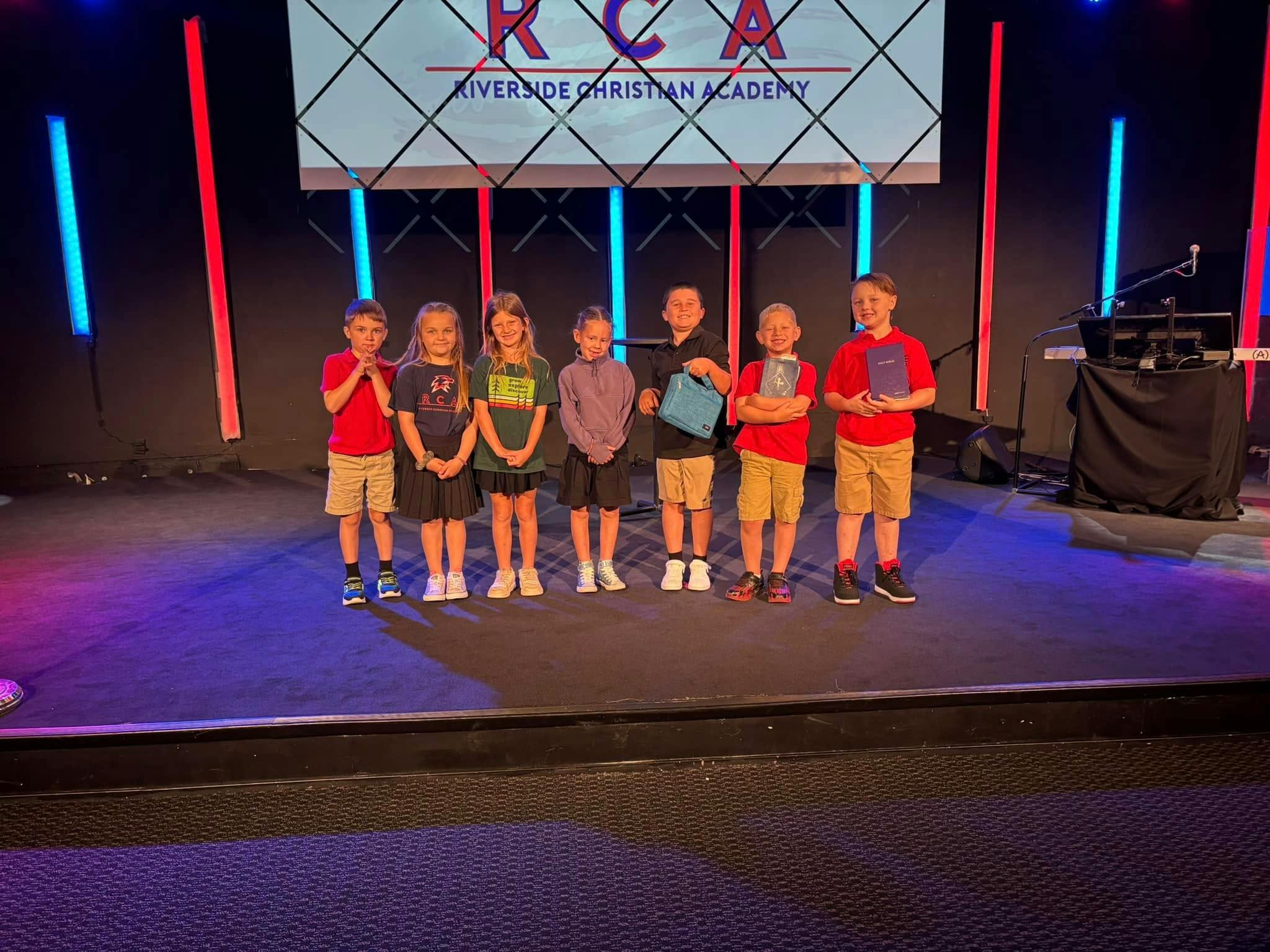 Six children standing on a stage with a screen displaying "RCA Riverside Christian Academy"