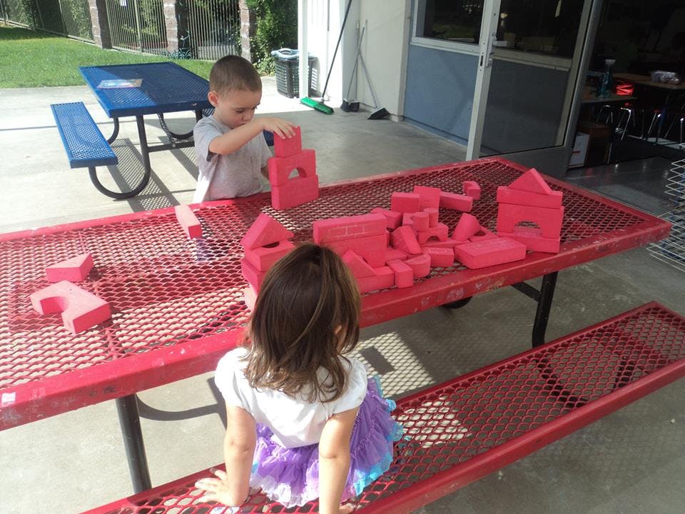 Child playing with red blocks on a picnic table while another child watches