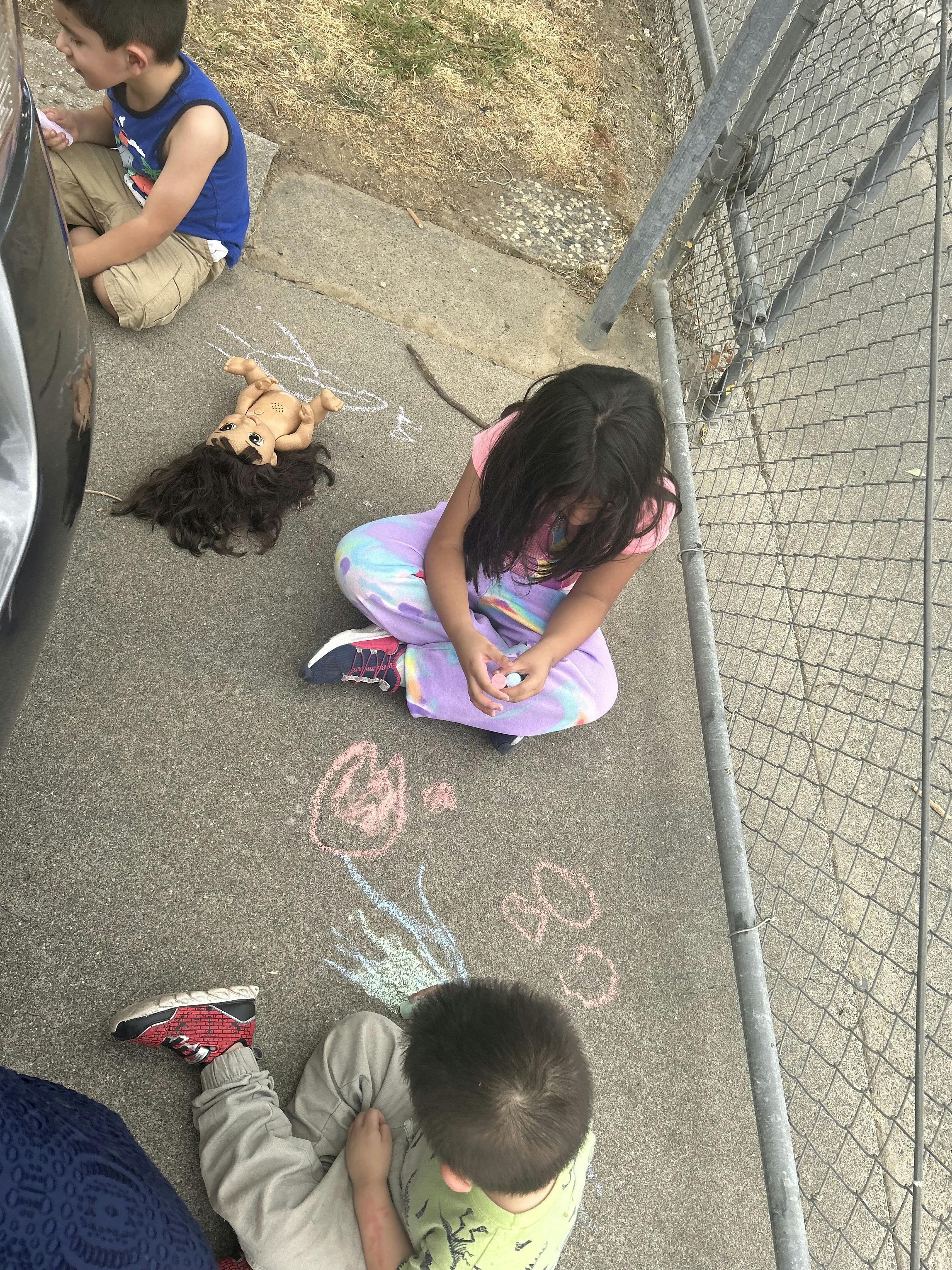 Children drawing with chalk on pavement next to a doll on the ground