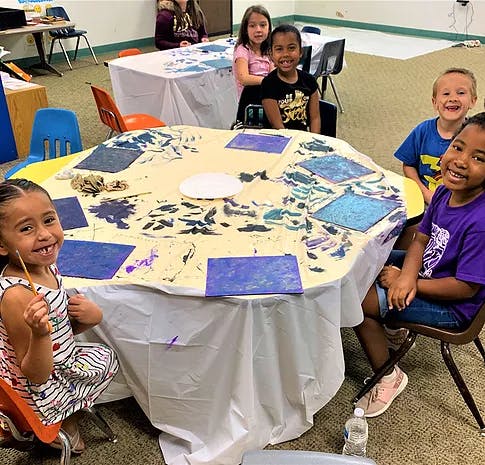 Children sitting around a table covered in paint and art supplies
