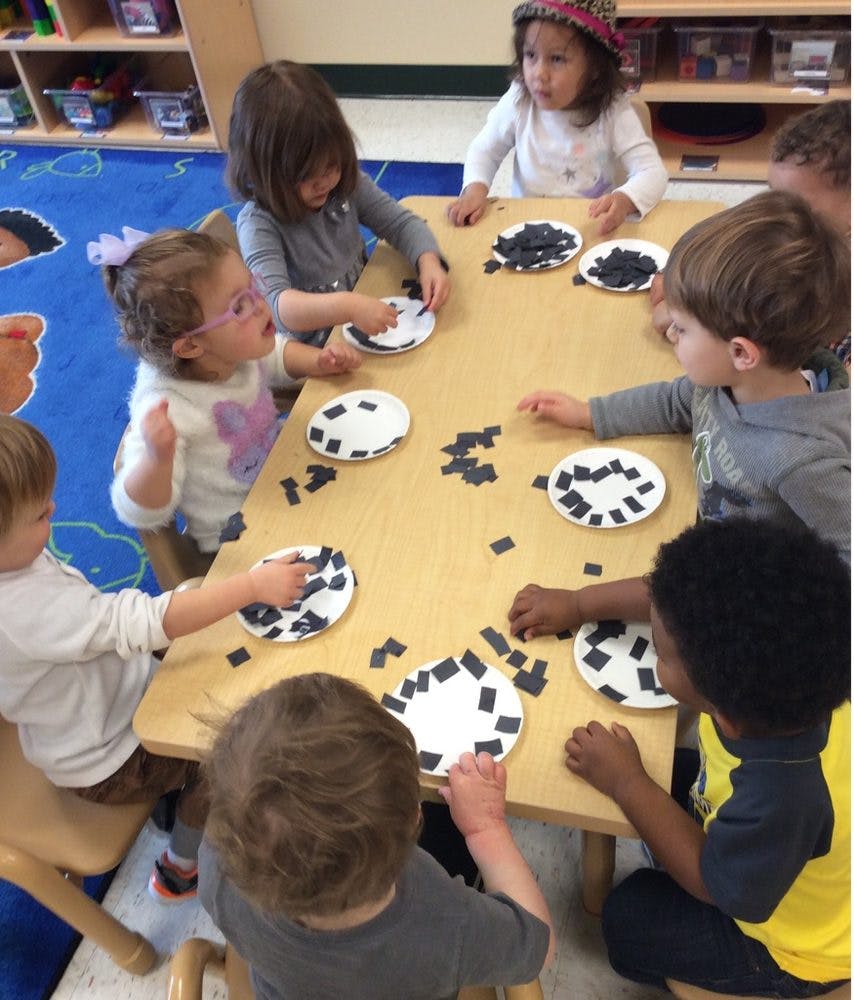Children at a table doing an art activity with paper plates and small black squares