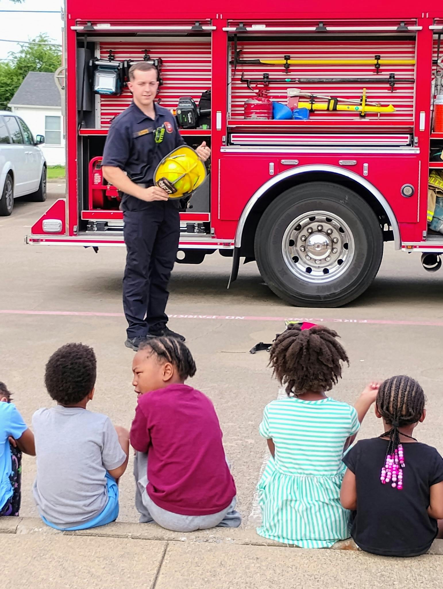 Firefighter holding a helmet, talking to children sitting on the ground