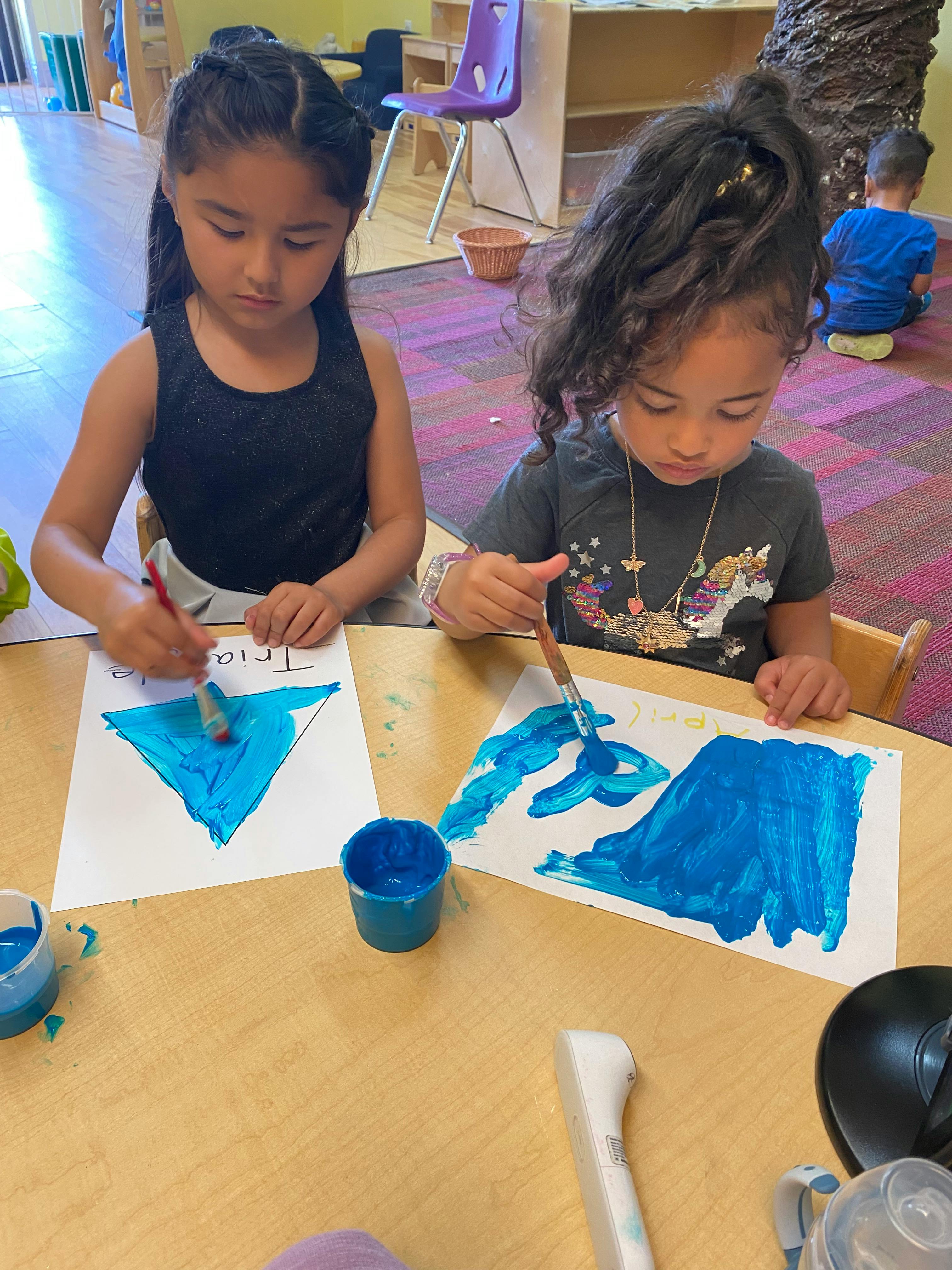 Two children painting shapes with blue paint at a table